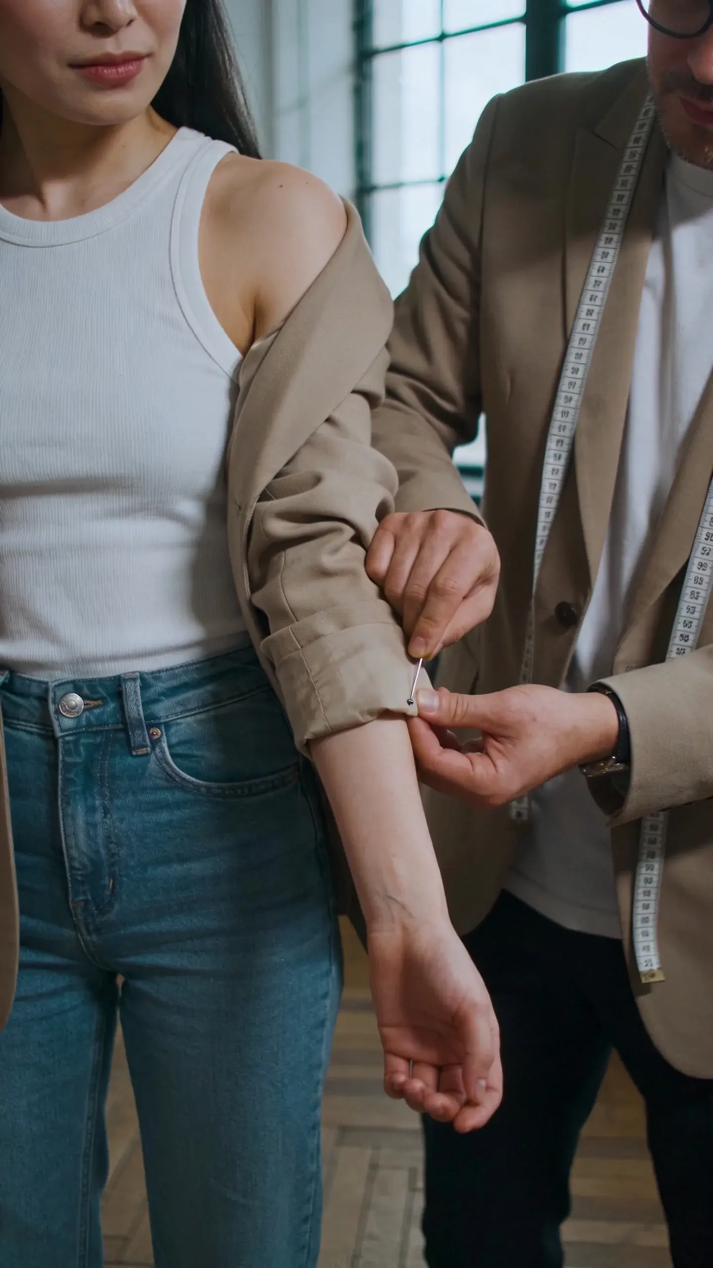 A boutique tailoring scene: a tailor pinning the sleeve length of a beige blazer on a woman wearing a white ribbed tank and straight blue jeans; measuring tape around the tailor’s neck; focus on the shoulder line and sleeve hitting just above the wrist bone; natural daylight through a window, wooden floor.
