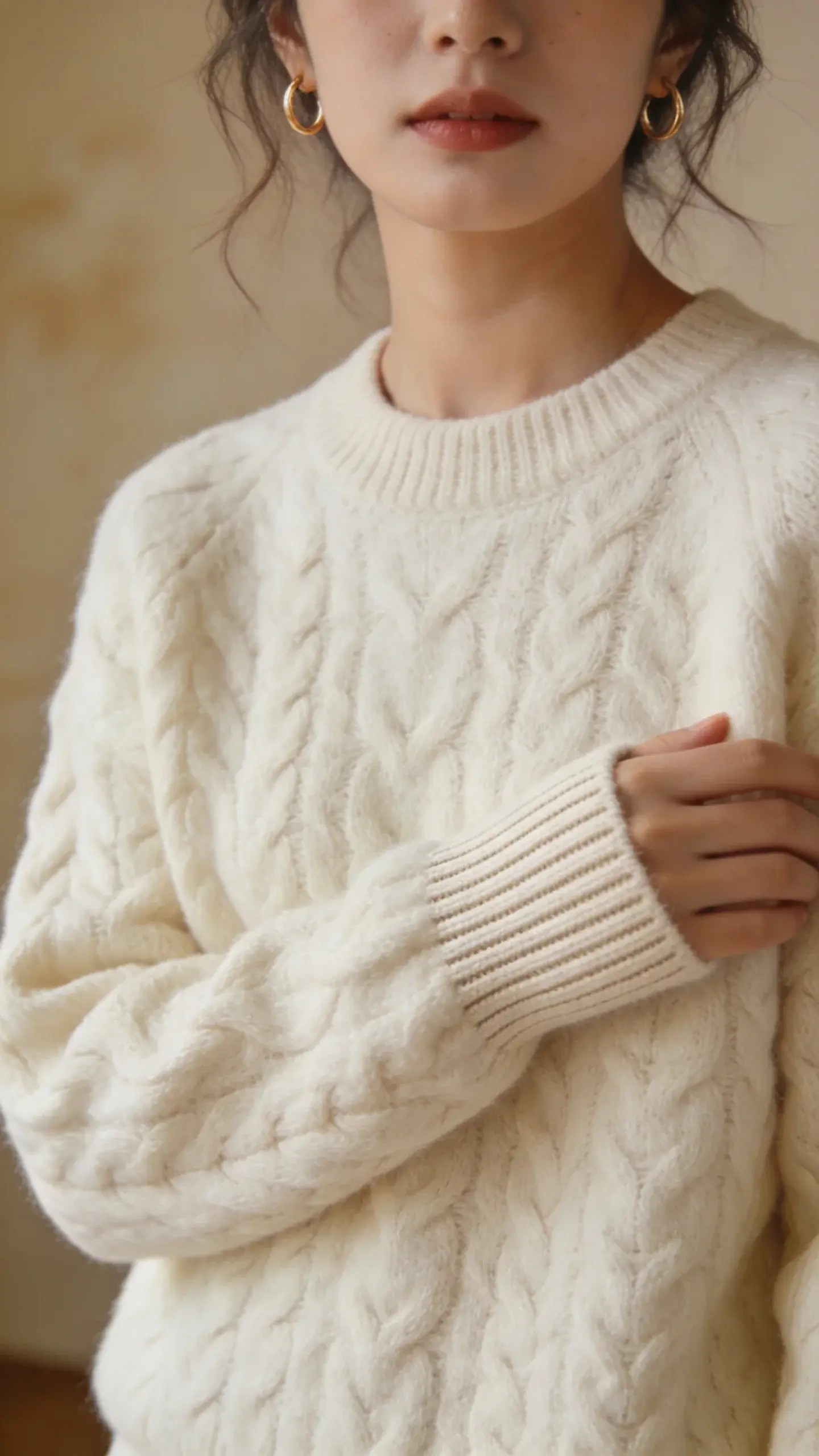 Knitwear focus: torso-up portrait of a woman wearing a chunky cable-knit cream sweater, fine-gauge ribbed cuffs, subtle gold hoop earrings; hair slightly tousled; background a softly blurred beige wall, showing richness and density of knit without any pilling.
