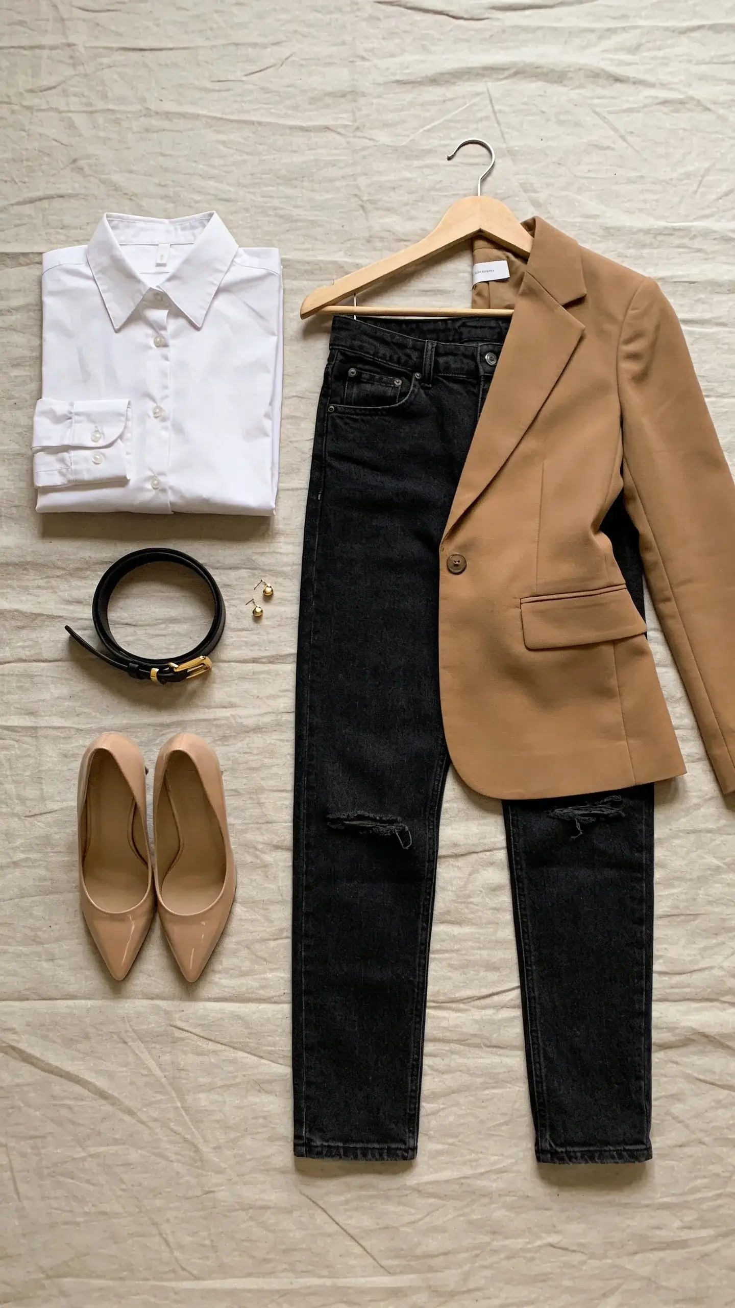 A minimalist wardrobe flat-lay on a neutral linen background: crisp white cotton poplin button-down neatly folded with visible collar and cuffs, slim dark-wash straight-leg jeans with no distressing, a camel single-breasted blazer with slightly nipped waist on a wooden hanger, a pair of polished nude leather pumps, a thin black leather belt with gold buckle, and delicate gold stud earrings; soft natural daylight, no text, clean editorial style.