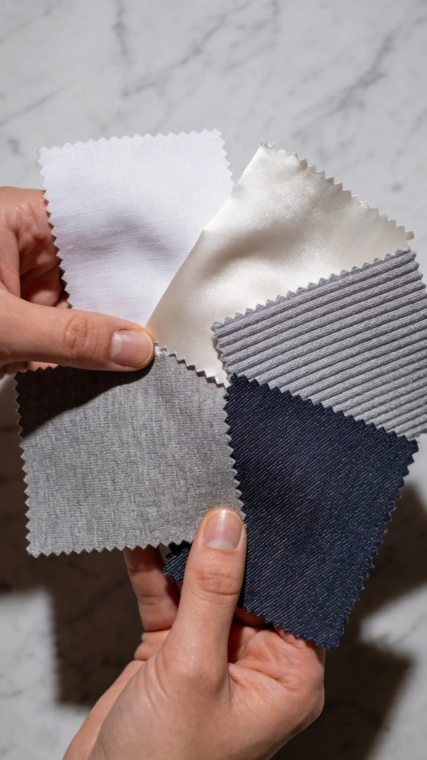 Close-up fabric study: hands gently holding and comparing swatches—cotton poplin, cotton-silk blend, ribbed knit, smooth knit, and dark denim—each labeled by texture through visual differentiation only (no text), arranged on a marble surface with soft shadows, showcasing quality weave and drape.