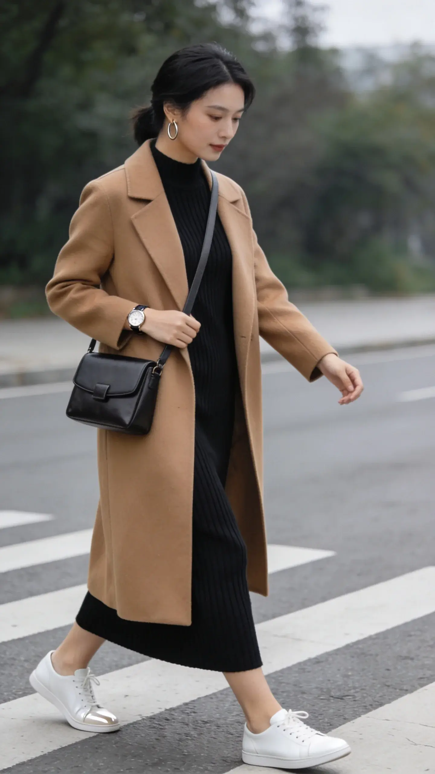 Street-style mid-shot of a woman in a ribbed black knit midi dress under a long camel coat, paired with sleek white low-profile leather sneakers and a structured crossbody bag; simple hoop earrings, subtle watch; she’s crossing a quiet crosswalk with elegant posture; overcast soft lighting, timeless vibe.