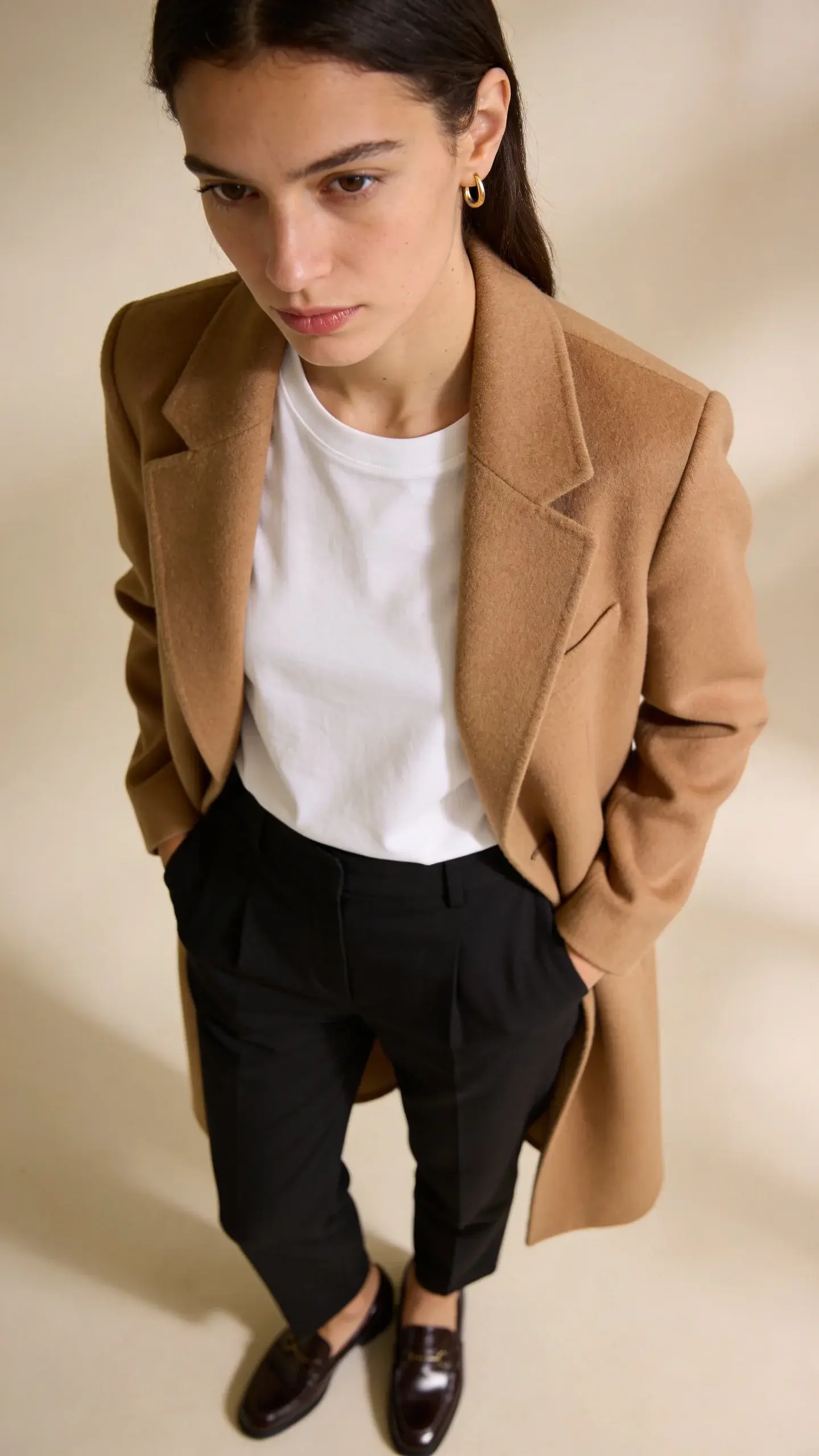 A minimalist, softly lit studio shot of a woman in a perfectly tailored camel wool coat over a crisp white crewneck tee and high-waisted black trousers, hands in pockets, wearing polished loafers and tiny gold hoop earrings; neutral backdrop, natural shadows, focus on fabric texture and clean lines, no logos.