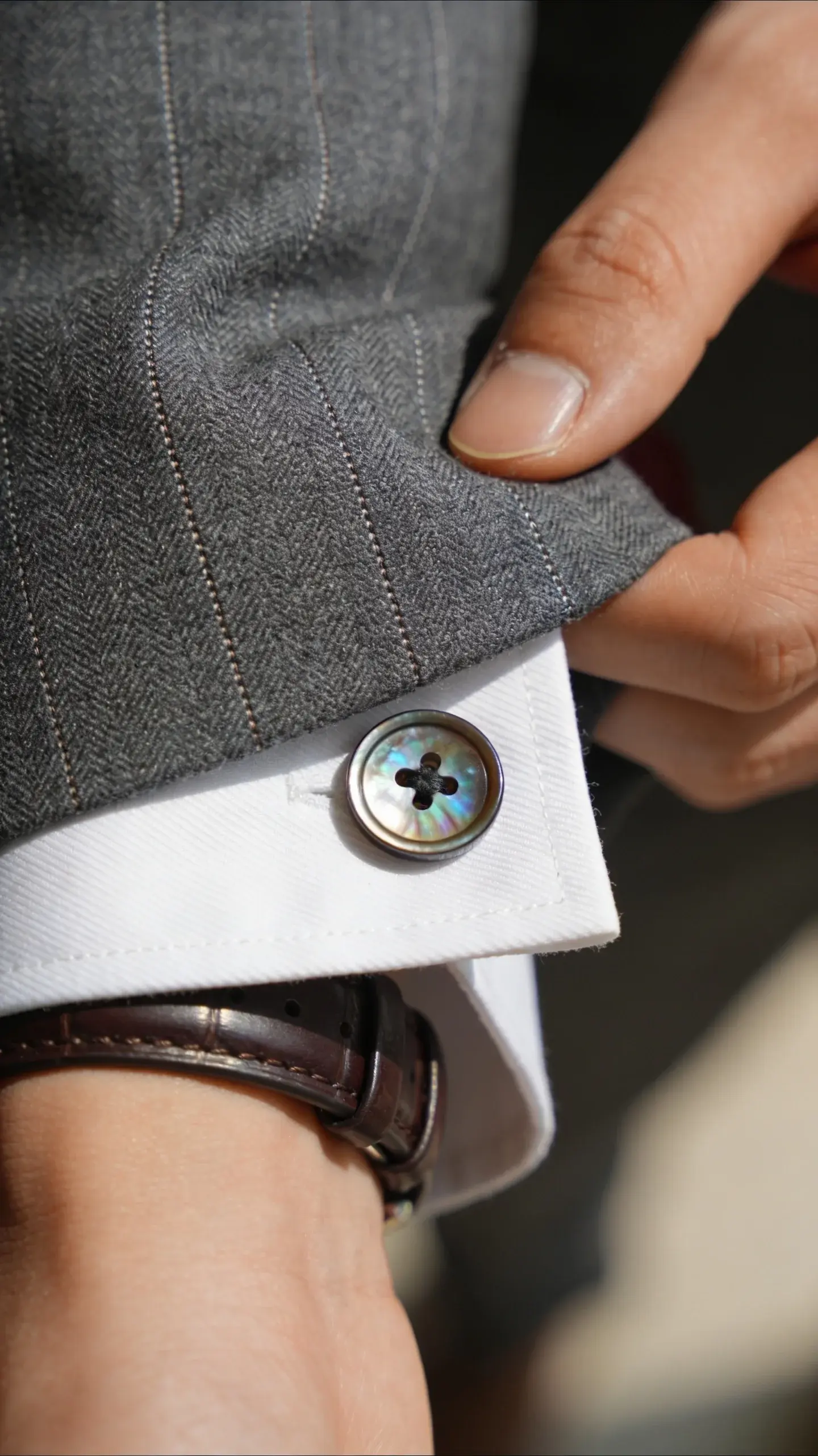 Detail shot of premium textures: a hand adjusting the cuff of a charcoal gray blazer with subtle herringbone weave, showing precise stitching, mother-of-pearl button on a white poplin shirt, and a thin leather watch strap in dark brown; shallow depth of field to highlight finish and fit.