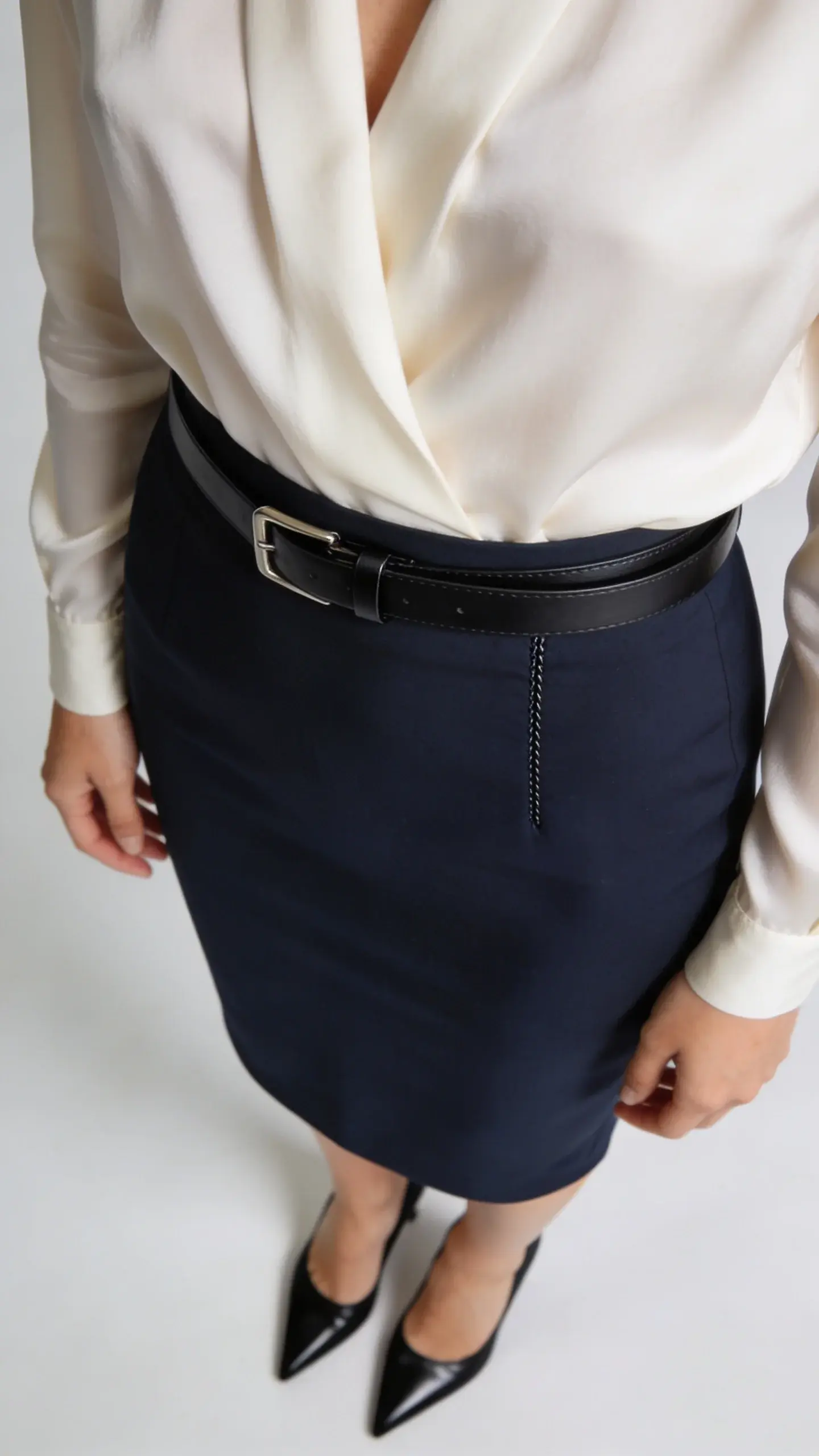 Workwear refinement: a close-up of a perfectly fitted navy pencil skirt paired with a tucked-in cream silk blouse, slim black leather belt with an unobtrusive buckle, and pointed black pumps; the blouse’s drape and skirt’s seam detail in crisp focus.