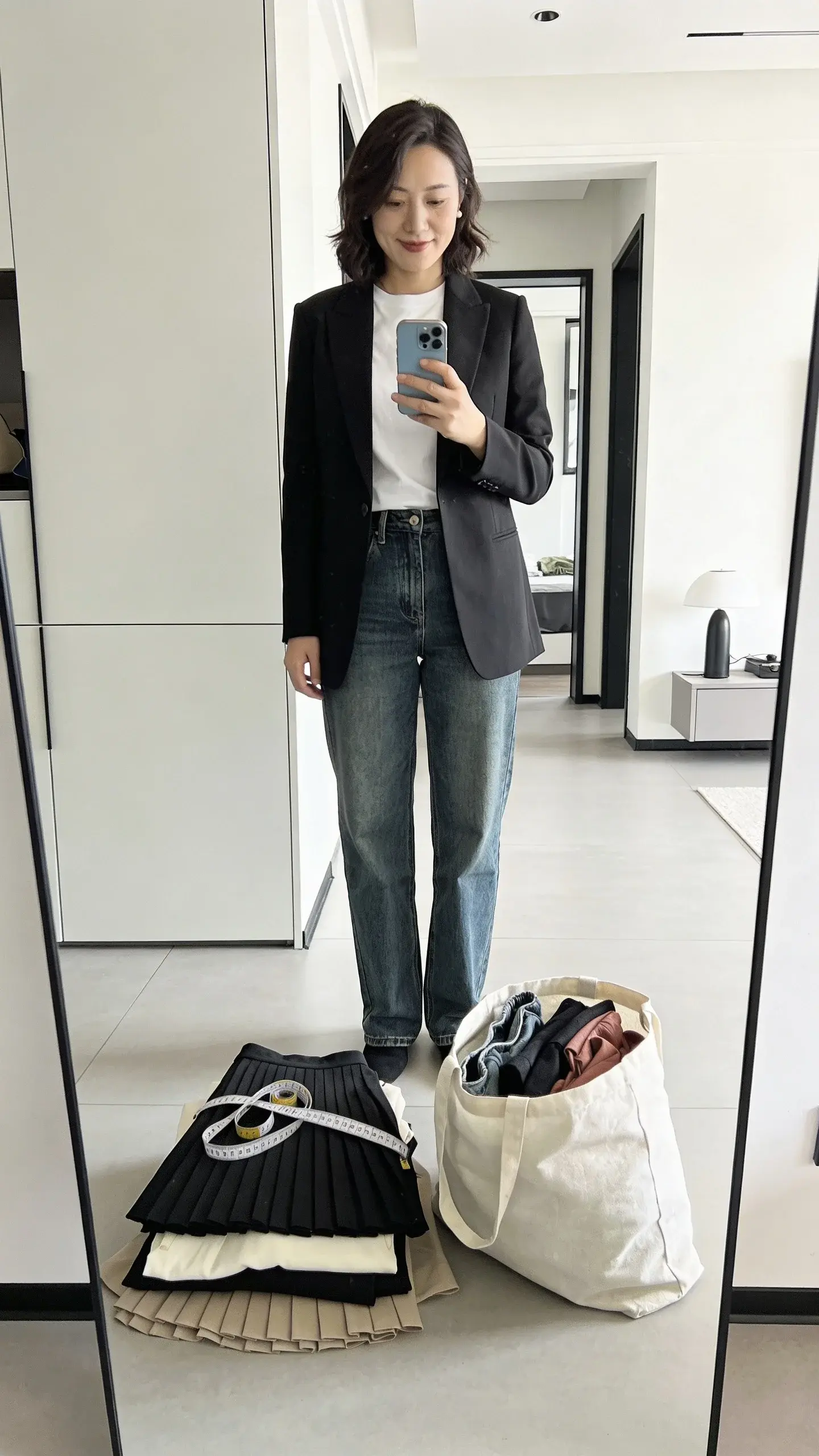 Chic woman in her 30s standing in front of a mirror taking a smartphone photo while trying on a tailored black blazer over a white tee and straight-leg jeans; two labeled piles visible on the floor by context: a “tailor” pile with a skirt and dress folded neatly with measuring tape on top, and a “donate” pile in a reusable bag; bright, clean modern apartment, no text.