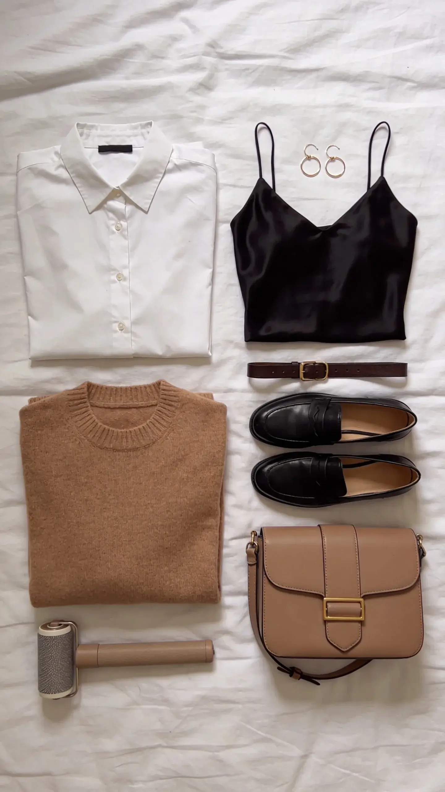 Overhead flat lay on a white bed of “polished essentials”: pressed white button-down, black silk camisole, camel sweater, slim leather belt, delicate gold hoop earrings, classic black loafers, structured taupe handbag, and a lint roller; everything neatly arranged with breathing space, no text.