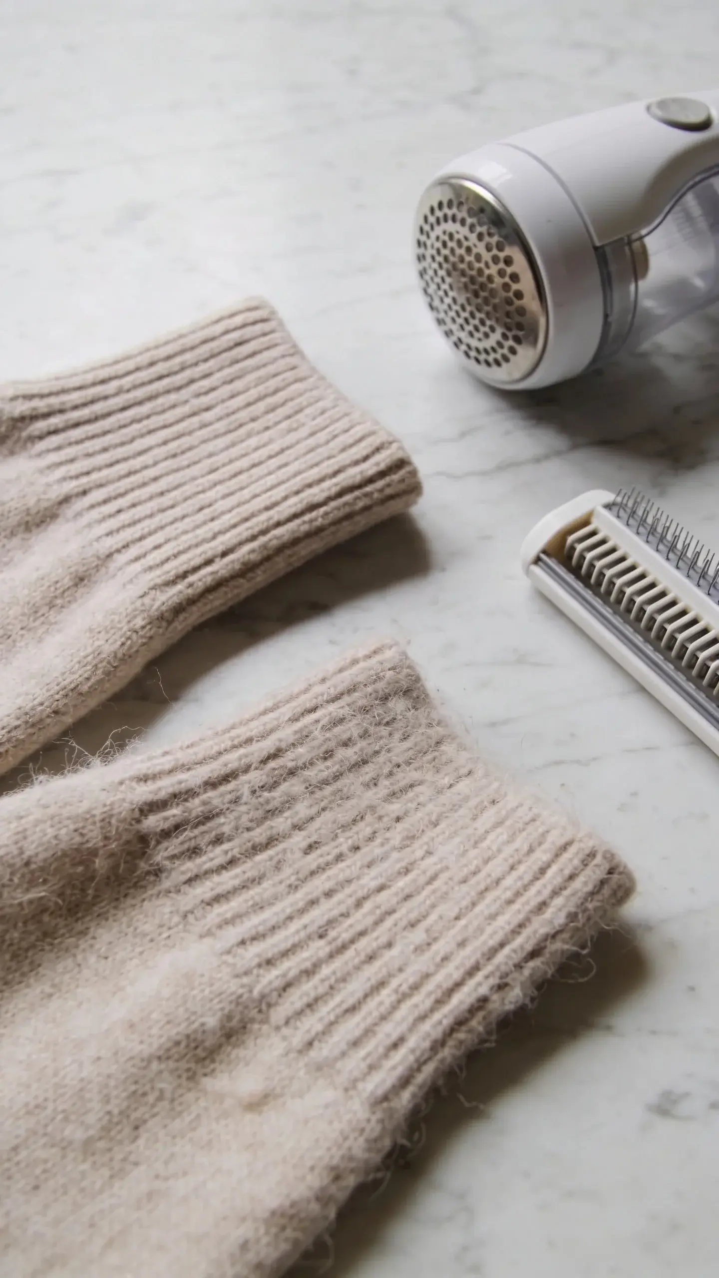 Detail shot of fabric quality: close-up comparison of a refined knit sweater sleeve (tightly woven, smooth) next to a pilled, loose-knit sleeve; a lint shaver and sweater comb nearby on a marble surface; calm, editorial lighting, no text.