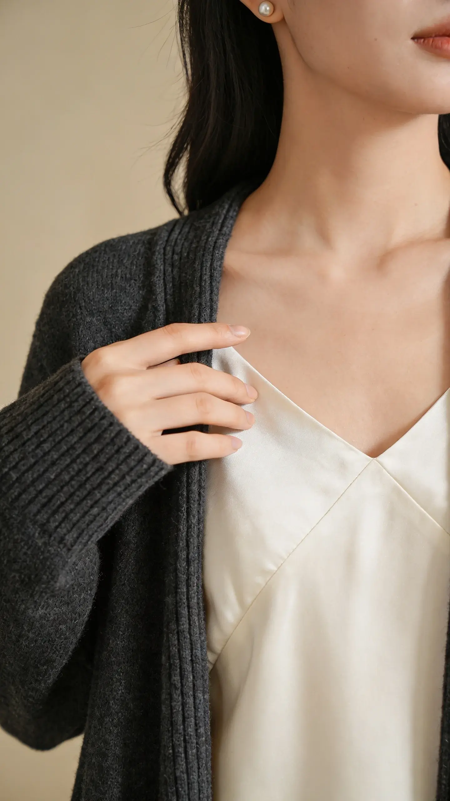 Close-up editorial shot focusing on fabric and finish: a hand gently skimming a cream silk slip dress under a charcoal cashmere cardigan; visible fine ribbing on the knit, bias-cut drape of silk, delicate pearl stud earring peeking from behind hair. Soft, diffused indoor light, warm beige background.
