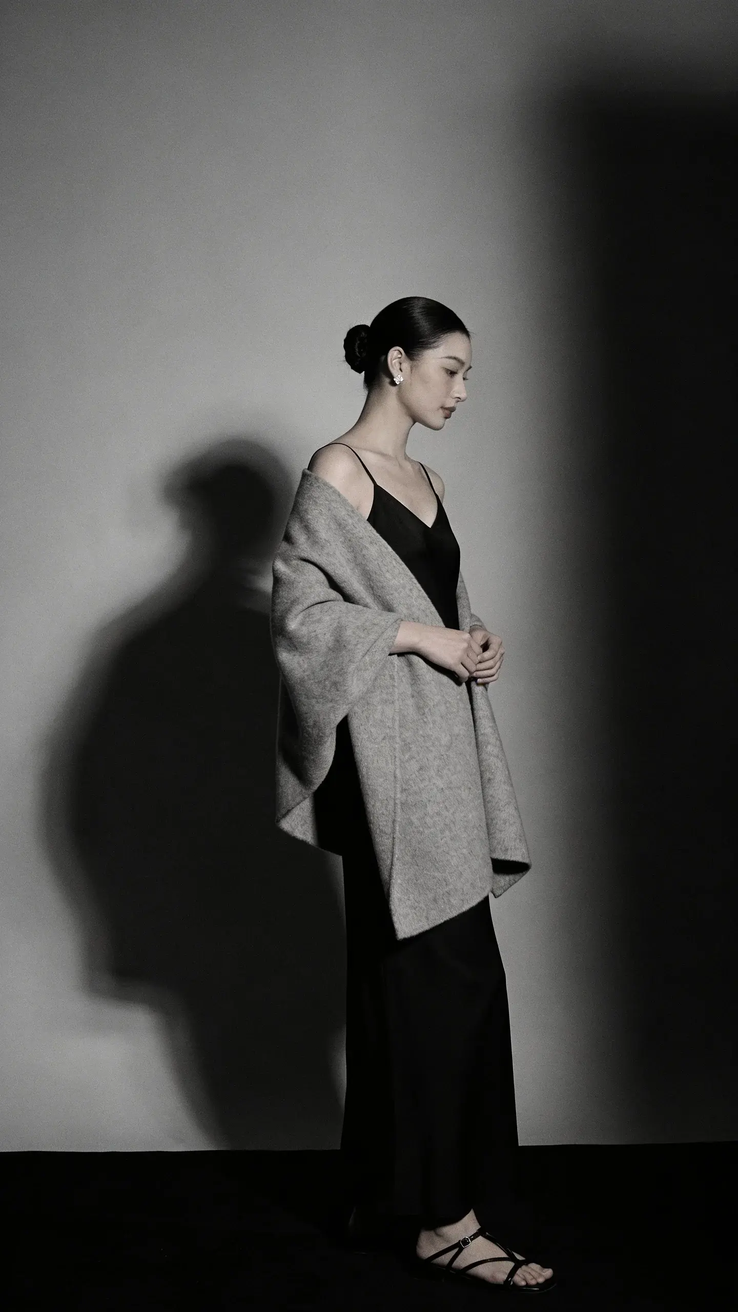 Evening minimalism: a floor-length black slip dress with clean neckline, draped under a light charcoal wool shawl; hair in a sleek chignon, small diamond studs, strappy black leather sandals. Low, moody lighting, soft shadows, no cluttered background.