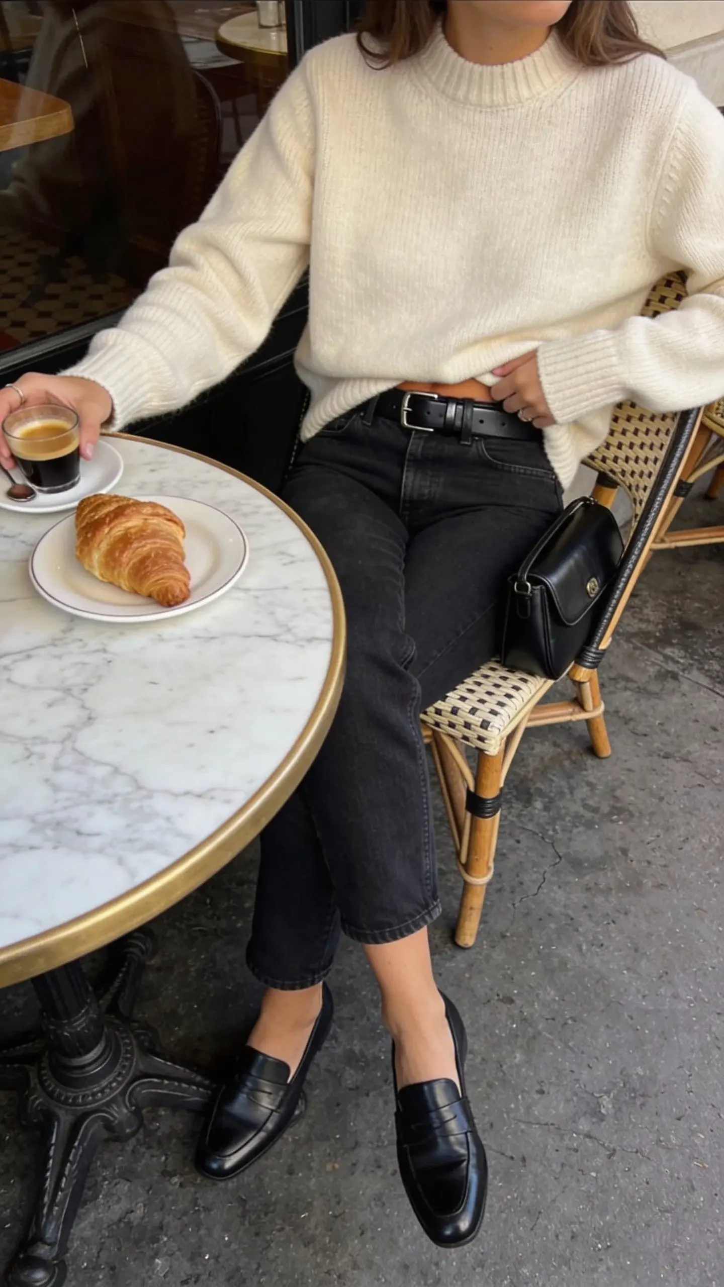 Elevated denim look: woman seated at a café table wearing a cream cashmere sweater half-tucked into dark straight-leg quality denim, black leather belt, polished black loafers; small structured black leather bag on chair. Soft morning café light, marble tabletop, croissant and espresso as props.