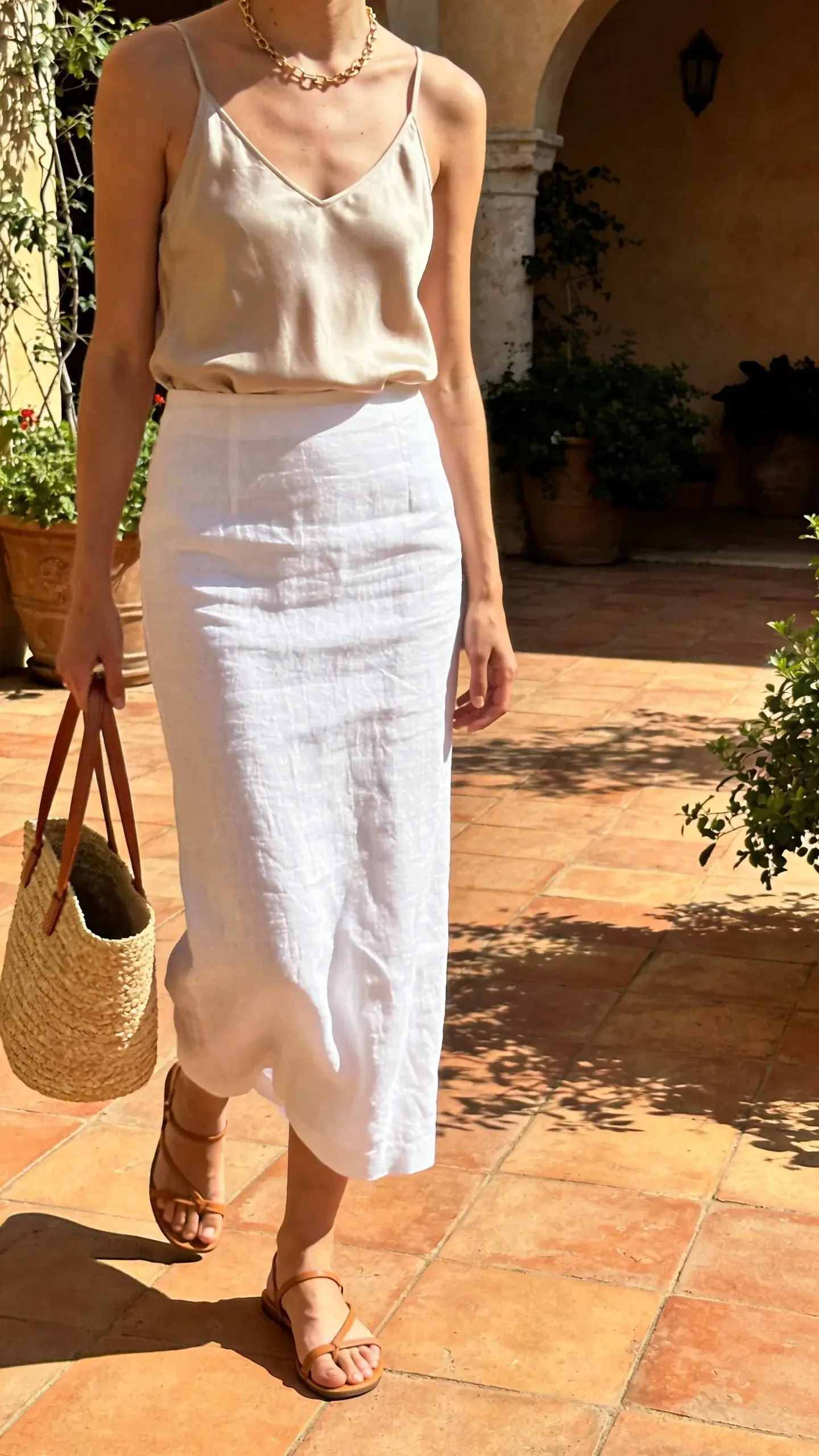 Summer quiet luxury: white linen-blend column skirt, sand-colored silk tank, thin tan leather sandals, straw tote with leather handles; delicate gold chain necklace. Sunlit Mediterranean-style courtyard, warm tones, breeze slightly moving the linen.