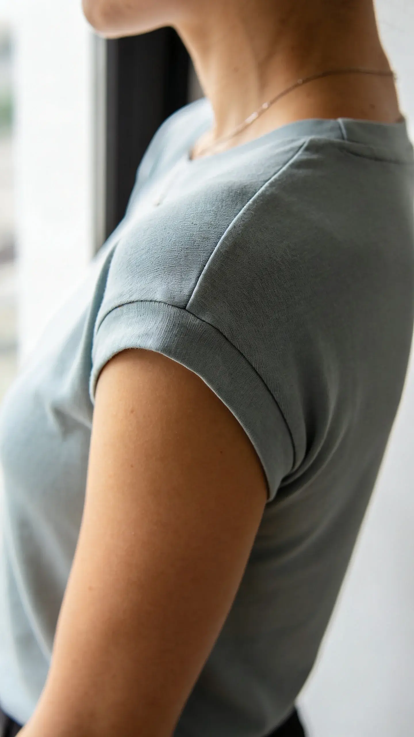 Close-up cropped shot of a woman’s upper torso focusing on perfect fit details: shoulder seam sitting at the edge of the shoulder, smooth tee fabric with no pulling, sleeves hitting mid-bicep, delicate necklace, soft daylight from a window, blurred minimalist interior background, high contrast but natural tones