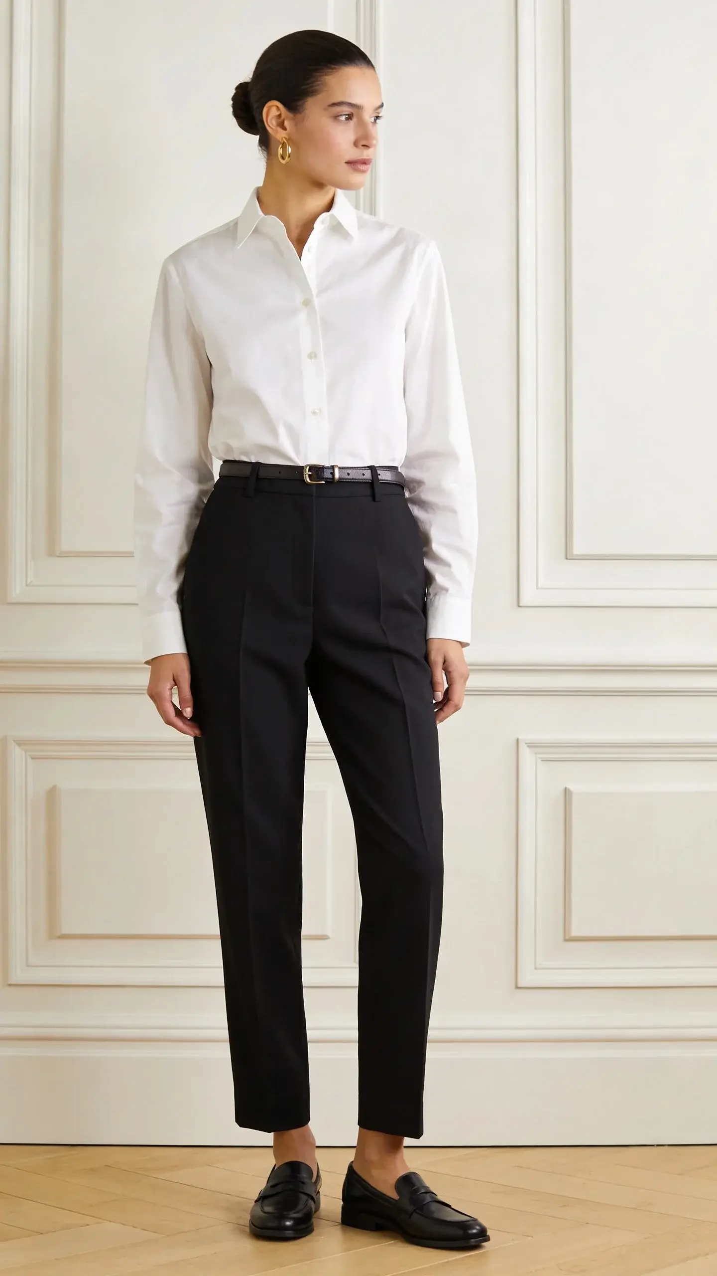 A minimalist, well-lit studio shot of a woman wearing a crisp white cotton poplin button-down tucked into high-waisted tailored black trousers, subtle gold hoop earrings, sleek low bun, leather belt, and polished black loafers; soft matte textures, no logos, neutral background.