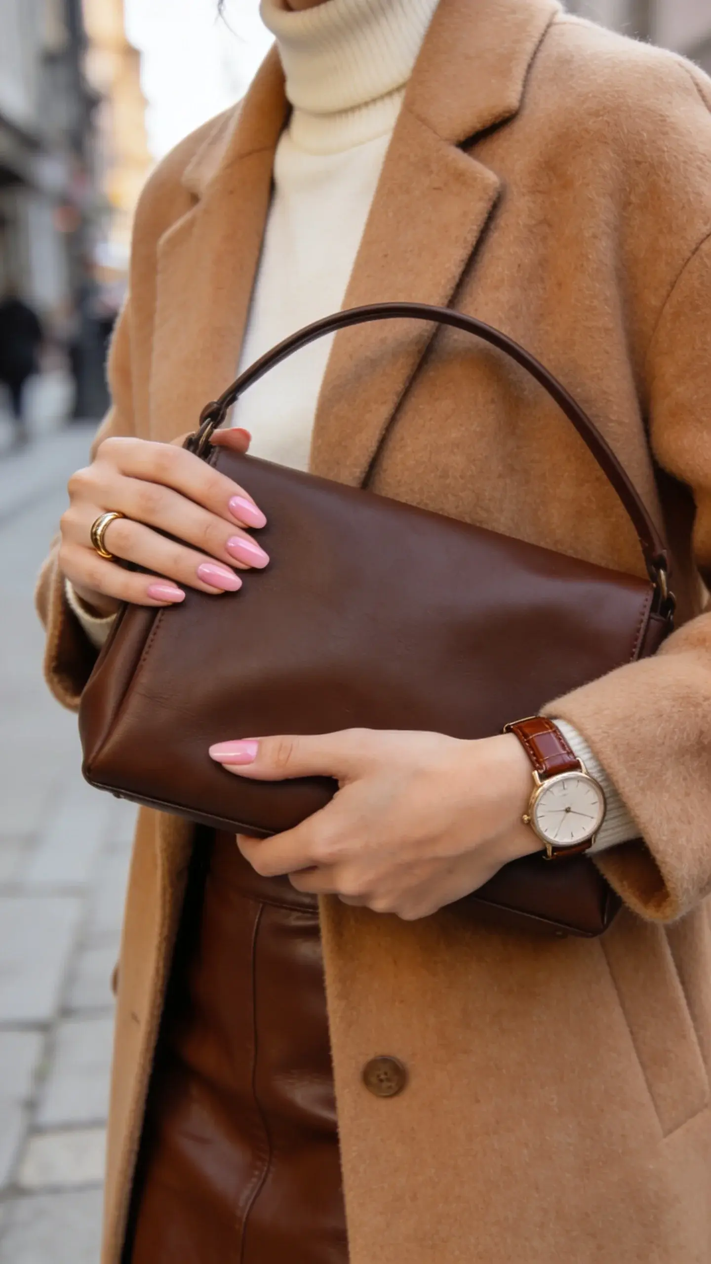 Street-style close-up of color harmony: warm camel wool coat over a cream turtleneck and chocolate leather skirt. Hands hold a rich brown structured handbag; visible details include neat manicure in sheer blush, simple gold ring, and refined watch with brown leather strap. Background is softly blurred city sidewalk.