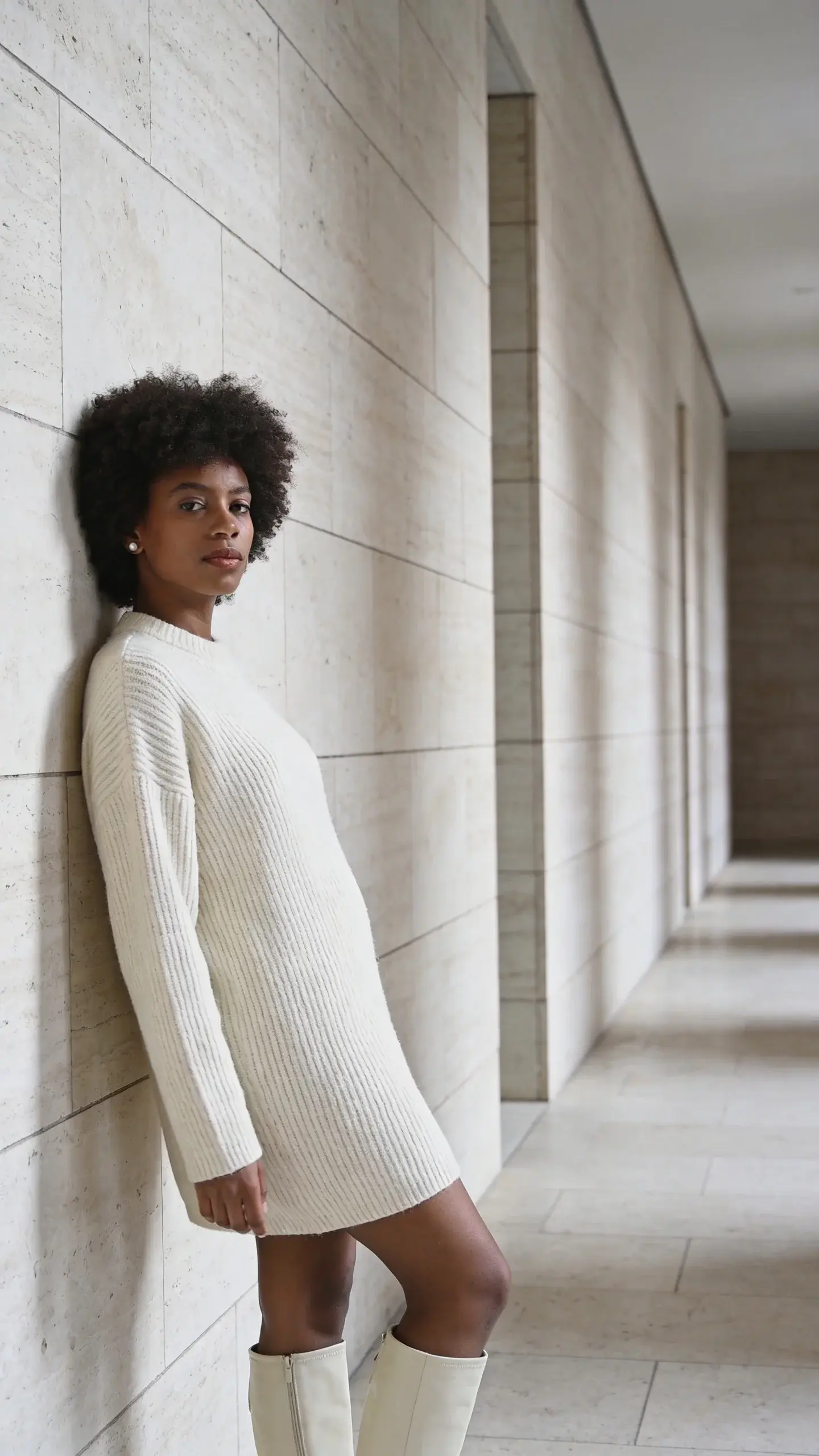 Minimalist hallway scene: a woman with natural curls and deep skin tone wears an all-ivory sweater dress with a ribbed texture, cream knee-high boots, and pearl stud earrings. She leans casually against a pale stone wall; soft diffused light, subtle shadows, serene mood.