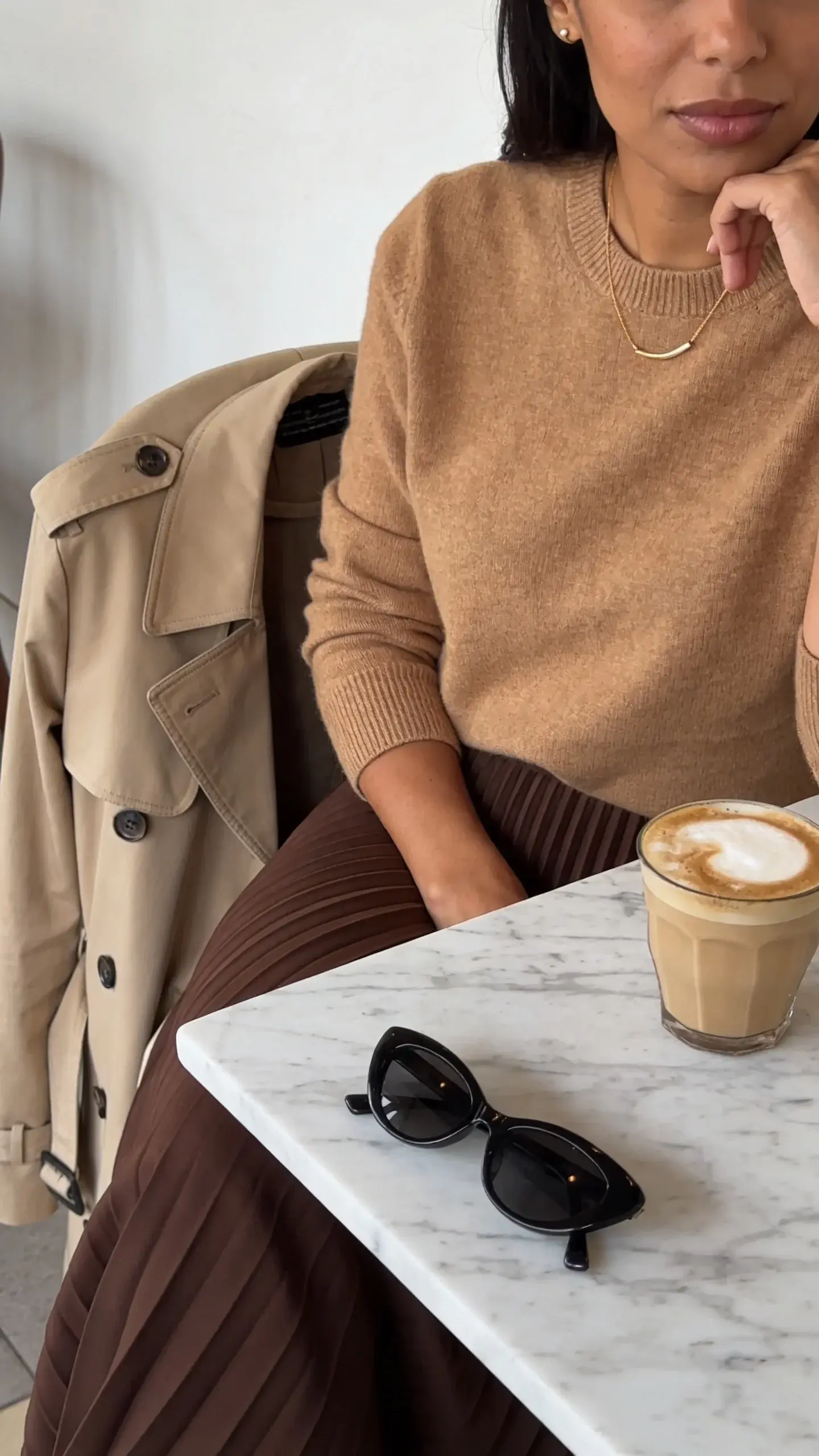 Elegant café moment: a woman with olive skin sits at a marble table in a camel cashmere crewneck, chocolate pleated midi skirt, and understated gold necklace. On the chair: a beige trench draped neatly; on the table: black sunglasses and a cappuccino. Background warm and minimal; no visible branding.