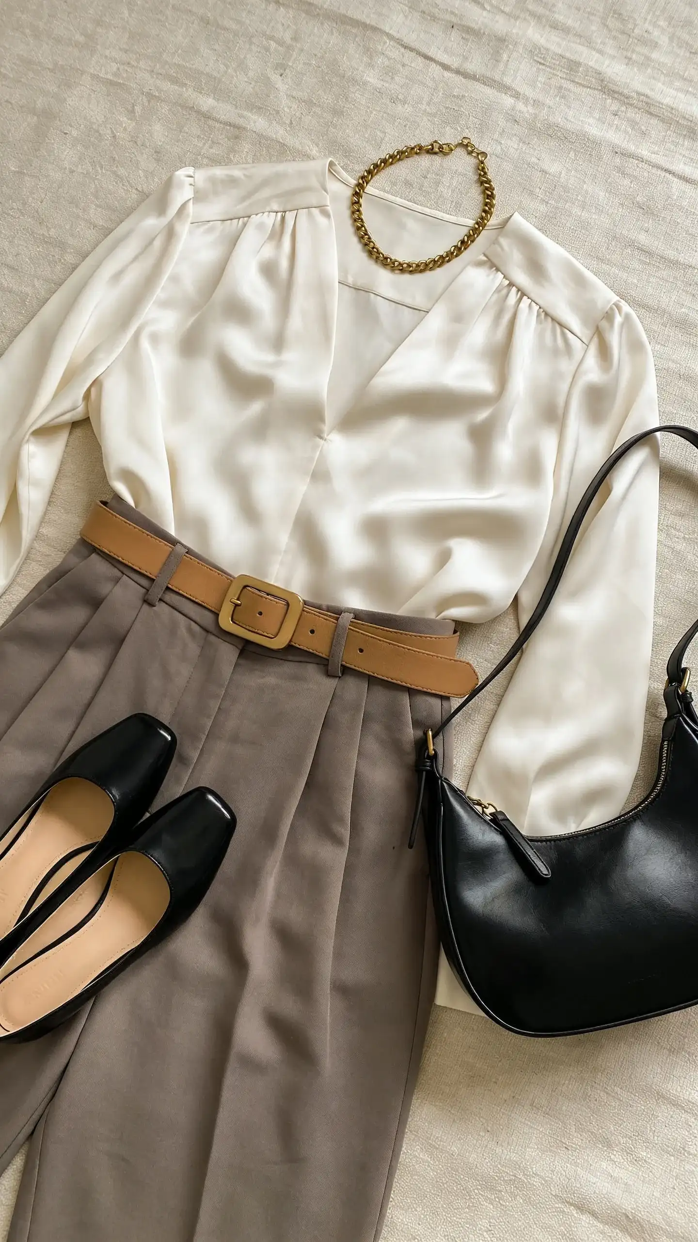 Flat lay of elevated neutral essentials: ivory silk blouse, taupe wide-leg trousers, camel belt with understated buckle, black leather pumps, delicate gold chain necklace, structured black handbag; arranged on a textured linen surface, soft overhead light.