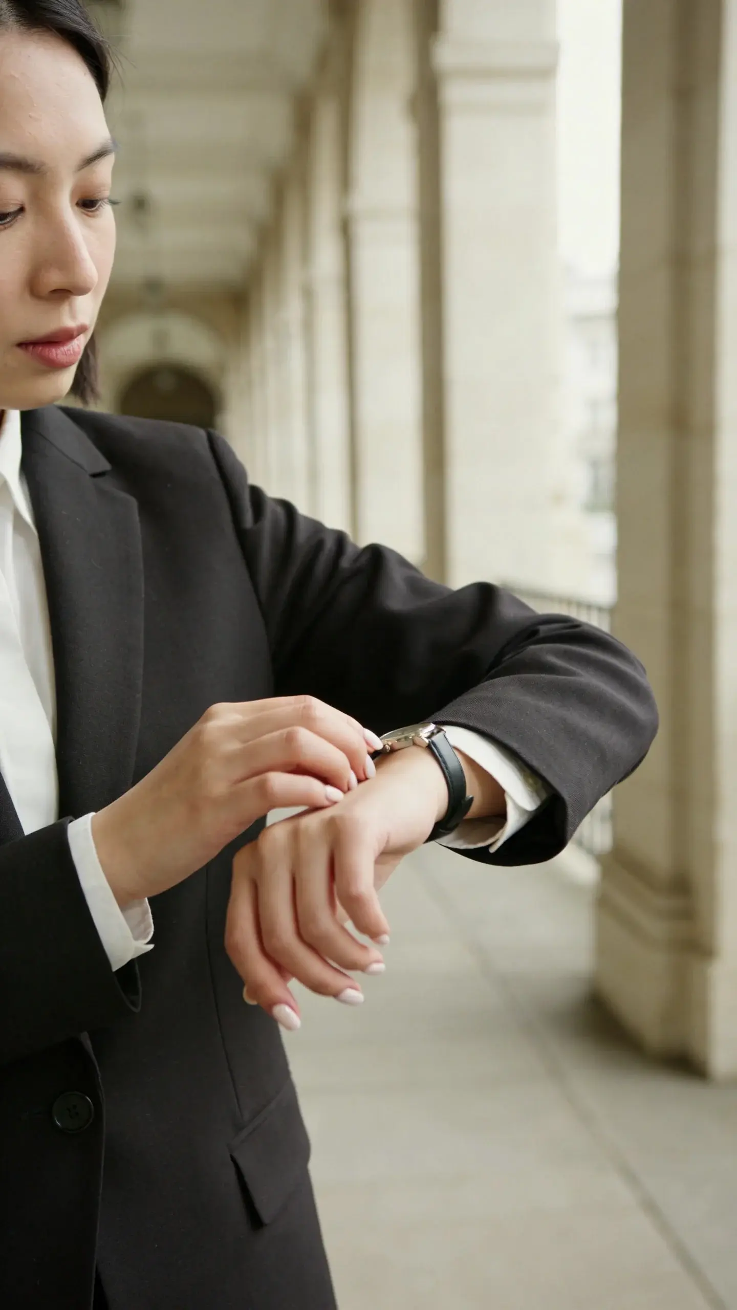 Minimalist hallway scene: a woman adjusting the cuff of her blazer to reveal a crisp shirt cuff and a slim watch; neat, short manicure; blazer fits neatly across the shoulders; soft side lighting, blurred architectural background.