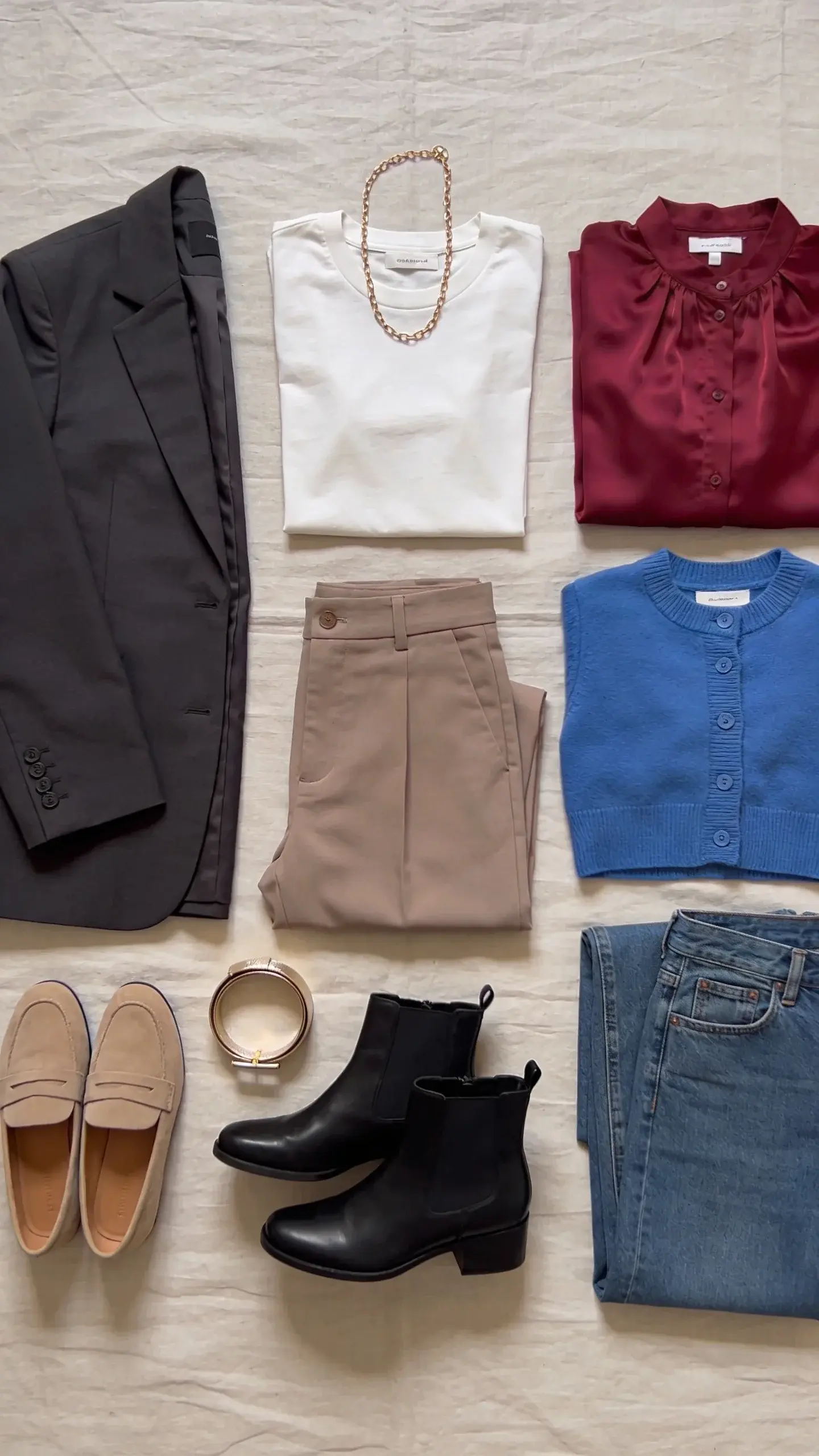 A neatly arranged flat lay of a women’s capsule wardrobe in a cohesive palette: base neutrals of charcoal, ivory, and taupe with accent pieces in burgundy and cornflower blue; include a charcoal blazer, ivory tee, taupe trousers, burgundy silk blouse, cornflower cardigan, classic straight-leg jeans, taupe loafers, black ankle boots, a slim gold chain necklace, and a champagne metallic belt; soft natural daylight, linen backdrop, no text.