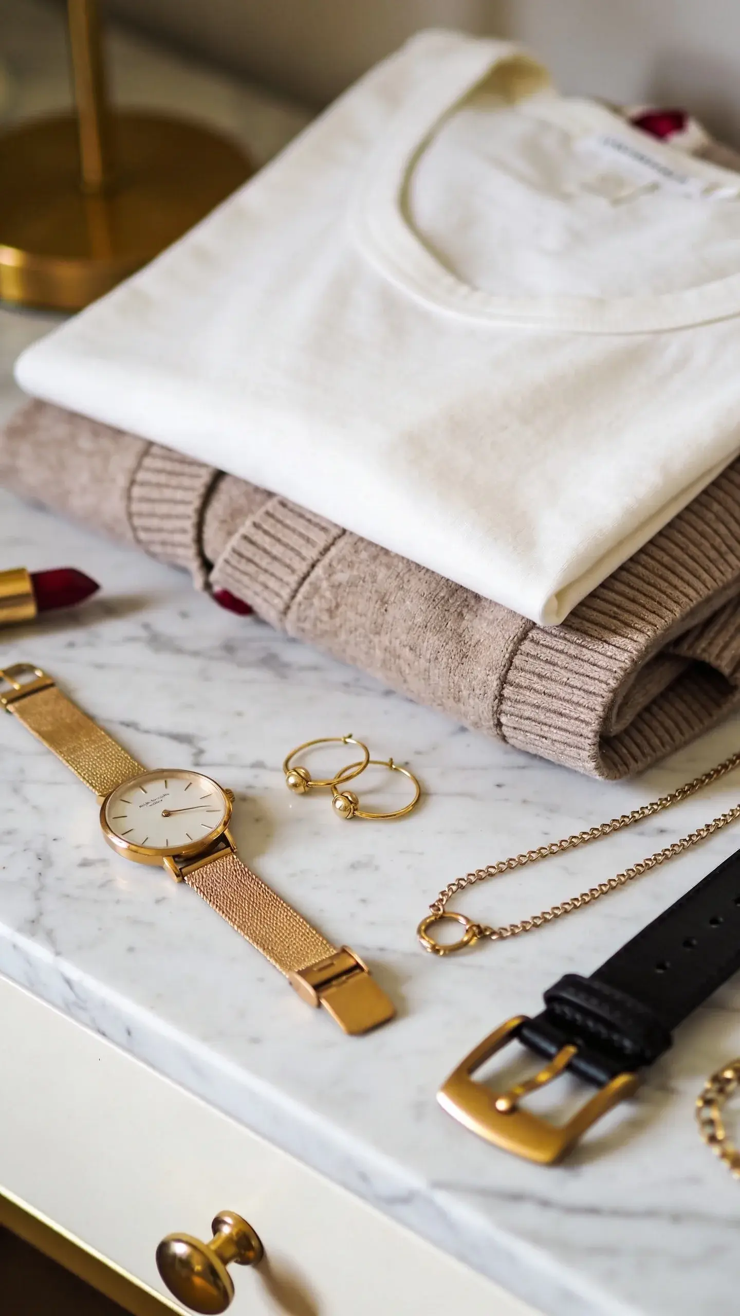Close-up styling vignette on a marble dresser: a curated selection of accessories in a single metallic—gold—featuring a slim watch, small hoops, delicate chain, and hardware of a black leather belt; beside them, folded ivory tee and taupe cardigan; a hint of burgundy lipstick; warm, diffused light.