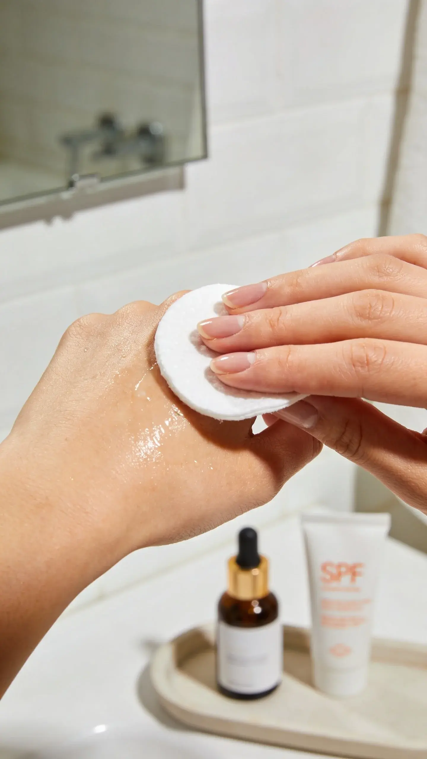 Close-up of a hand sweeping a cotton pad soaked with micellar water across a dewy cheek in front of a bathroom mirror; clean nails, soft morning light, blurred background showing a simple tray with just a serum bottle and SPF tube.