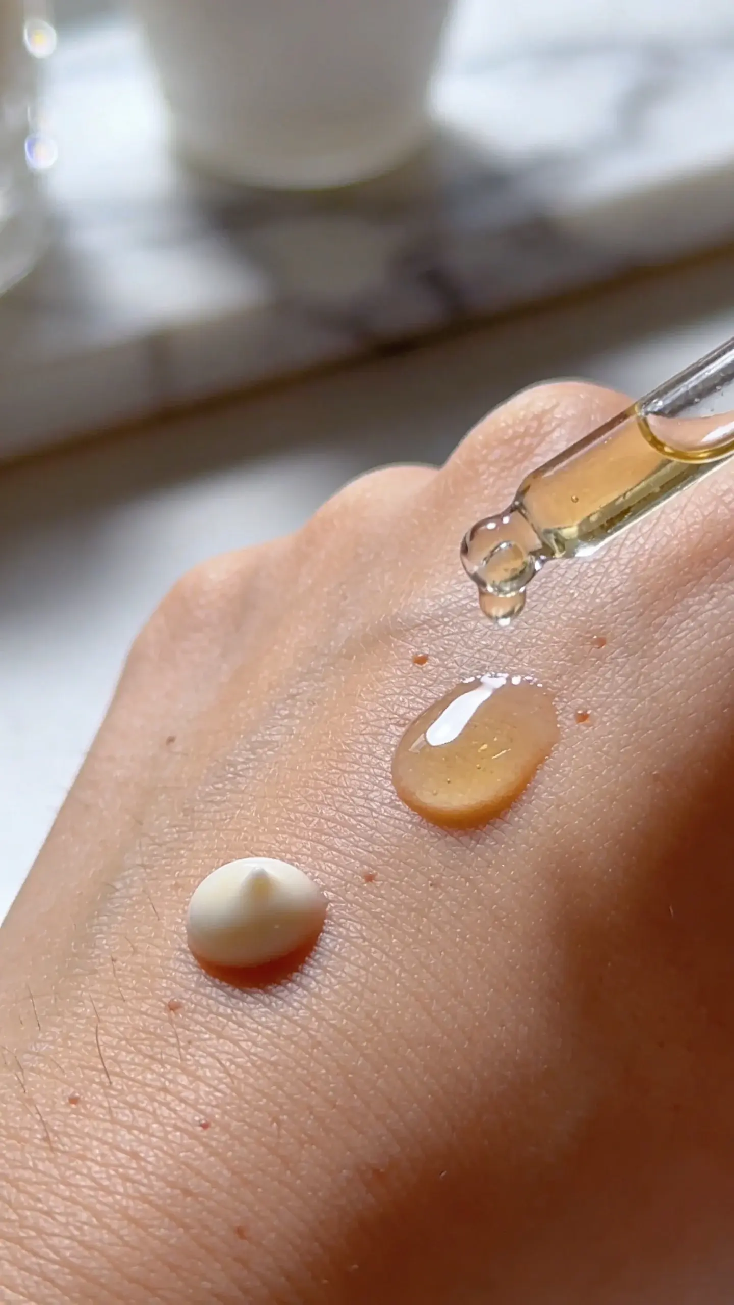Hyper-detailed macro shot of a single serum drop on the back of a hand next to a small pea-sized bead of sunscreen; skin texture glowy, balanced highlights, marble counter blurred in background.