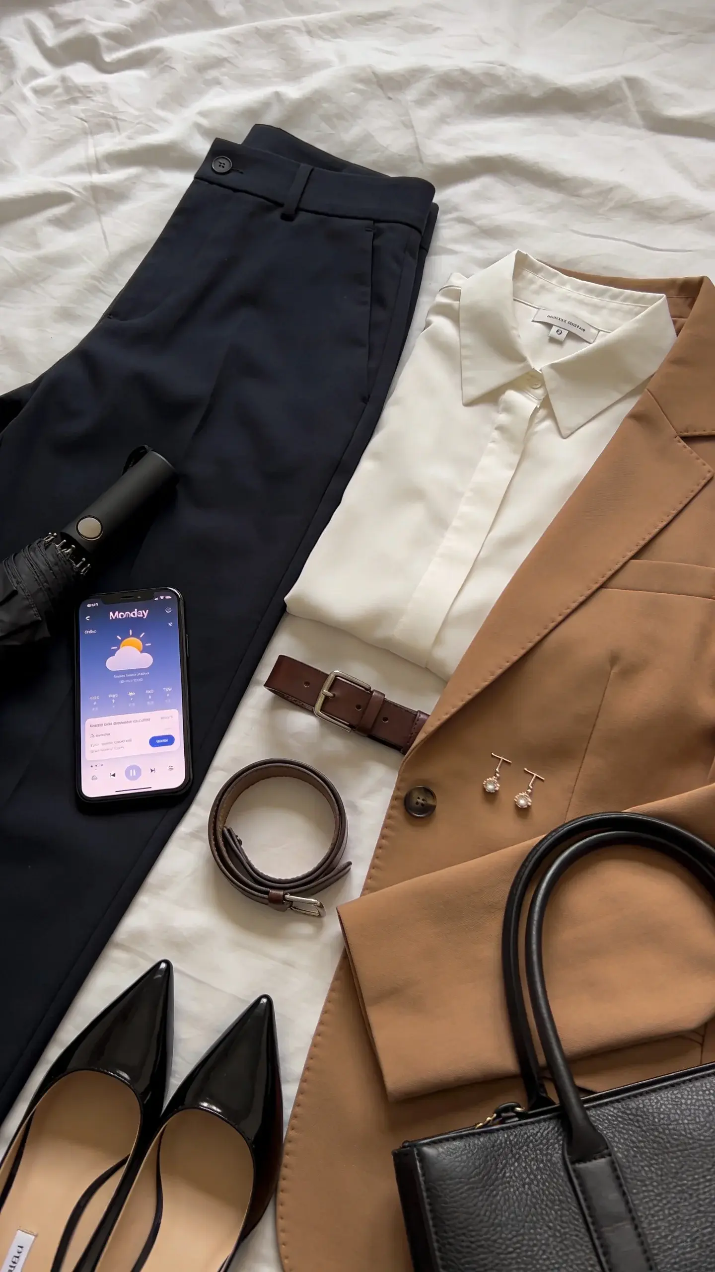 A Monday work outfit laid out on a bed: navy tailored trousers, crisp ivory blouse, camel blazer, slim leather belt, pointed-toe loafers, delicate stud earrings, and a structured black tote; a weather app on a phone beside a folded compact umbrella; serene, tidy composition, no text.