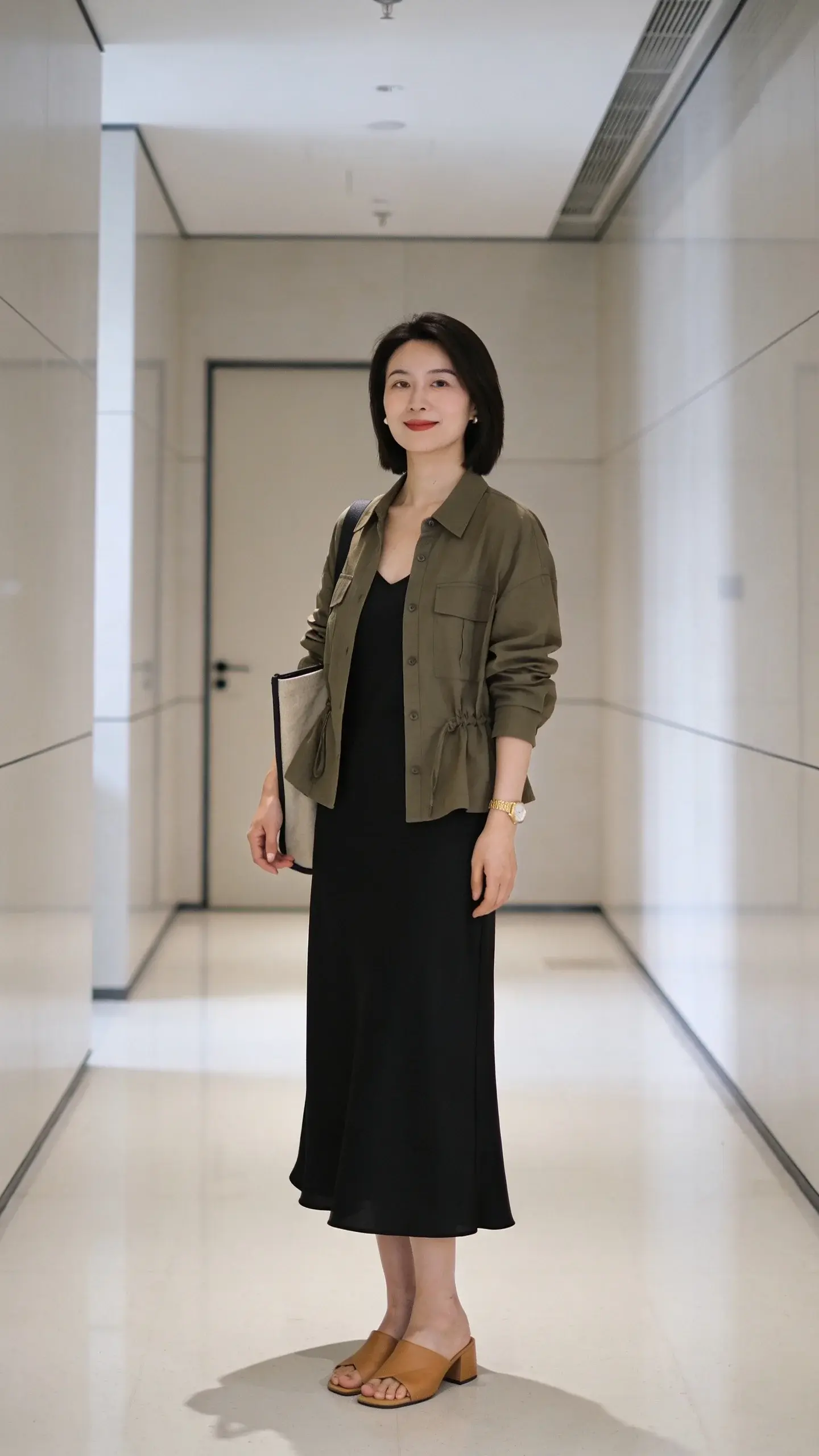 Office hallway setting: a woman in her 30s wearing a simple black midi slip dress, an olive utility jacket slightly cinched at the waist, tan low block-heel sandals, a minimalist gold watch as her signature accessory, and a tote bag; neutral background, modern aesthetic, no text.