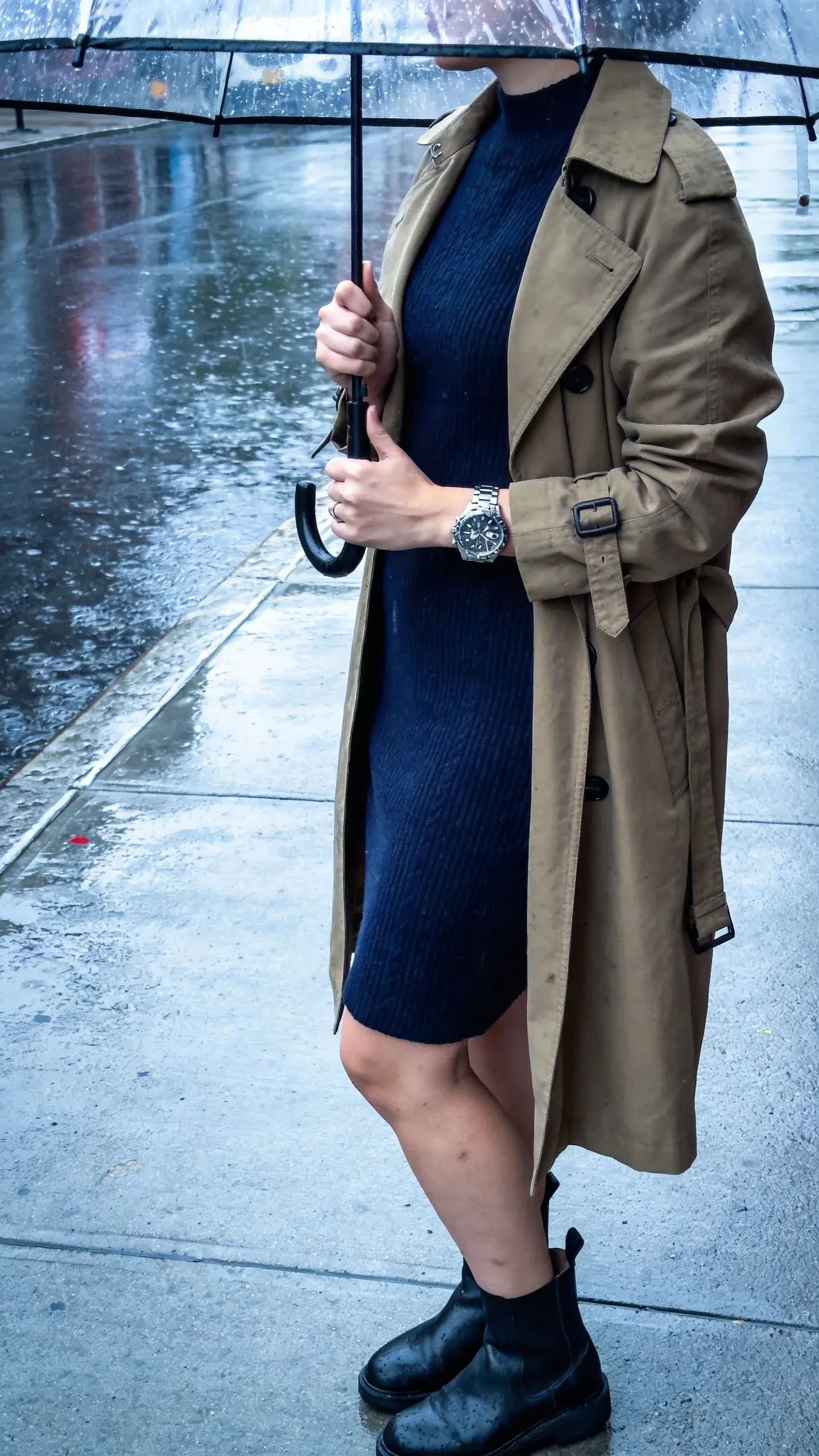 Rainy-day sidewalk: a woman wearing a navy knit dress (knee-length, short sleeves), a khaki trench coat, black Chelsea boots, and a chunky silver watch as the signature; holding a clear umbrella, slick pavement reflections, no text.