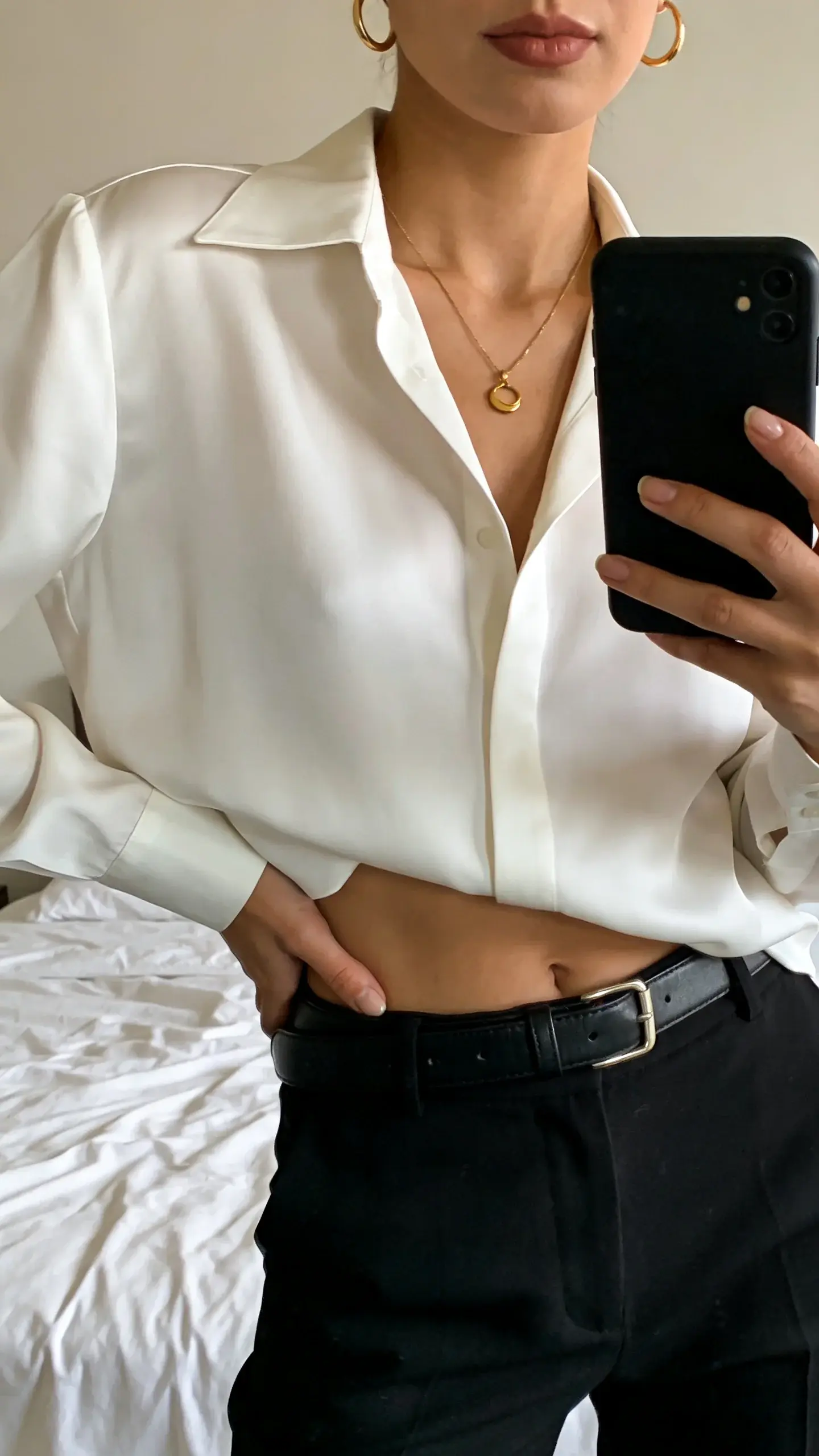 A close-up mirror selfie vibe (no visible face, cropped at shoulders): elegant woman in black high-waisted trousers, white silk blouse half-tucked, black leather belt, delicate gold necklace and hoops. She holds a phone with a simple black case; background is a neutral bedroom with crisp white bedding. Soft, flattering morning light.