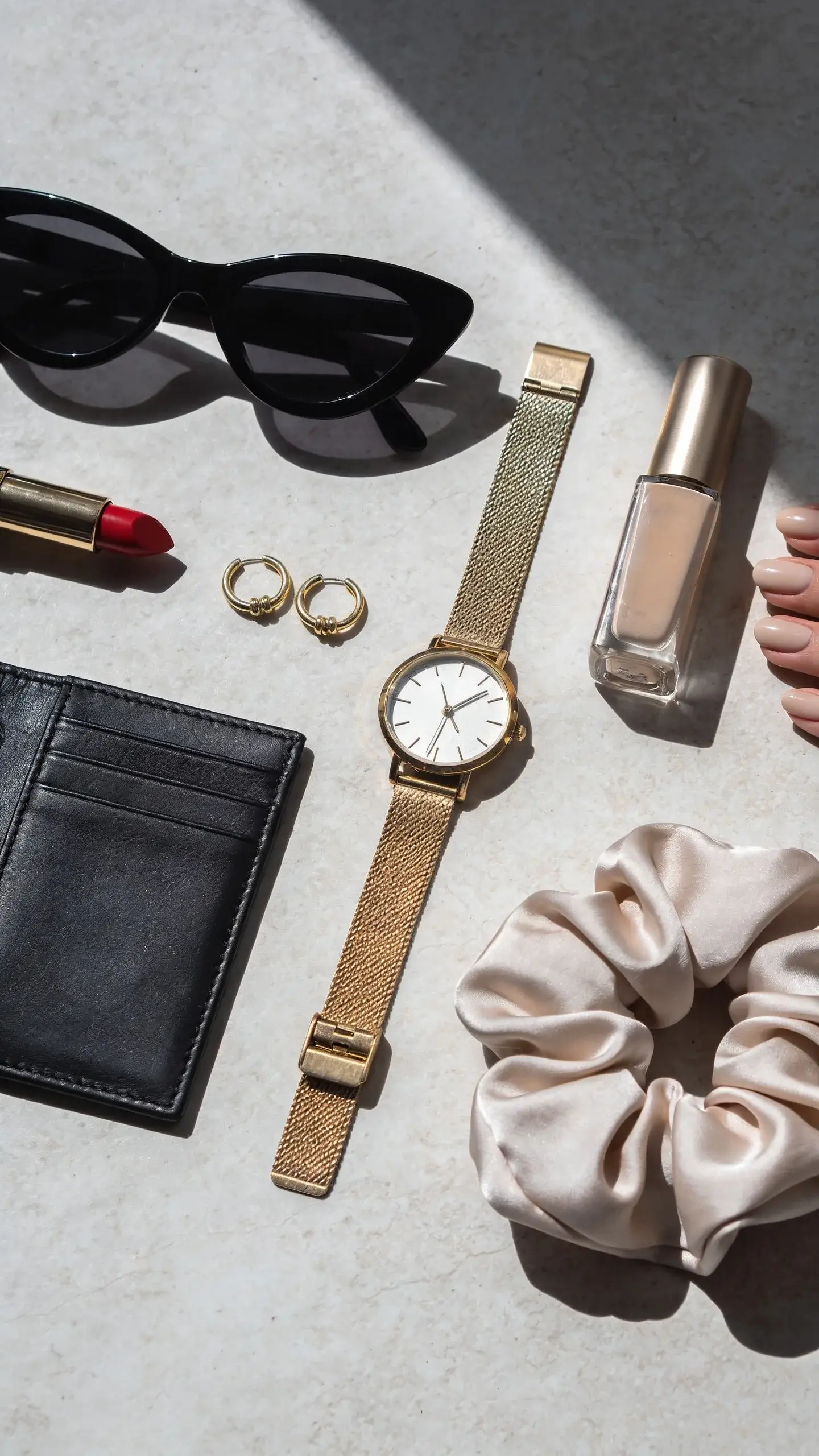 A beauty and accessory flat lay complementing the capsule: black cat-eye sunglasses, slim gold watch, small gold huggie earrings, a single red lipstick, a neutral nail polish, a silk hair scrunchie, and a black leather cardholder. Background is a light stone surface; soft shadows, refined and minimal.