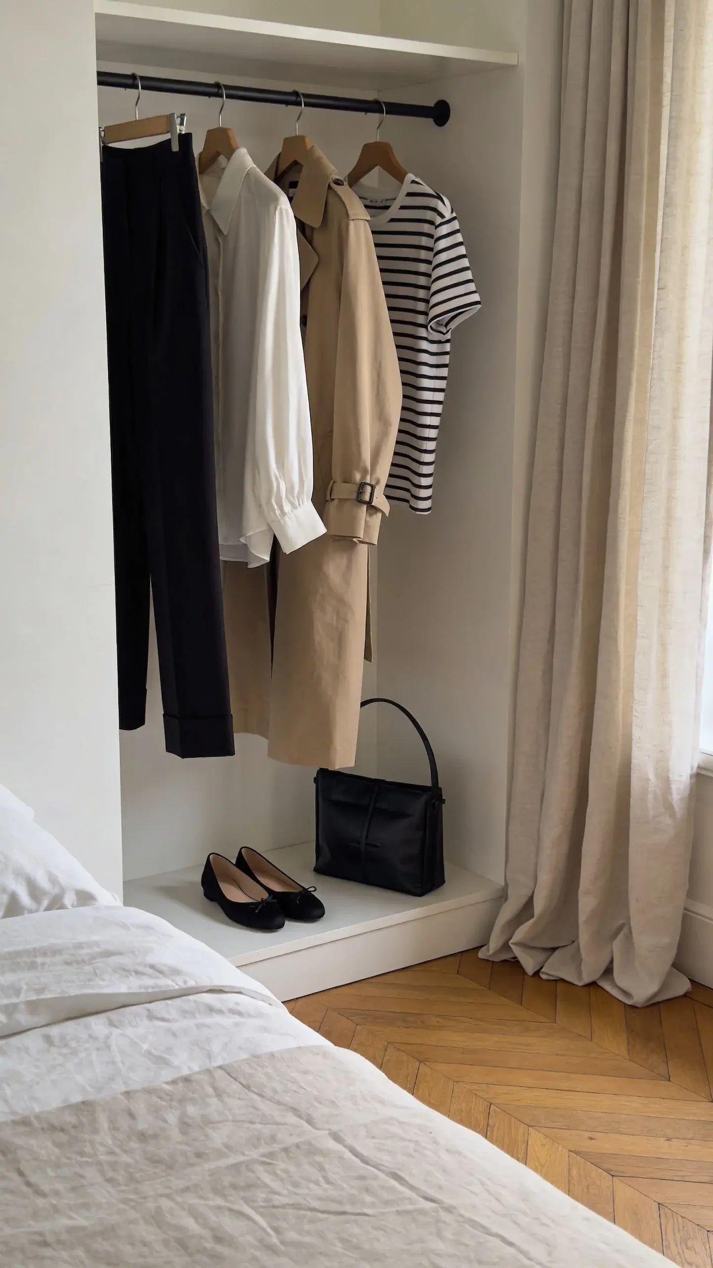 A serene bedroom corner with a minimalist open clothing rack featuring a tightly curated Parisian-minimal capsule: black cigarette trousers, white silk blouse, beige trench coat, striped Breton tee, black ballet flats, and a structured black bag; soft morning light, neutral palette, no text, clean styling, wooden floor, linen curtains.