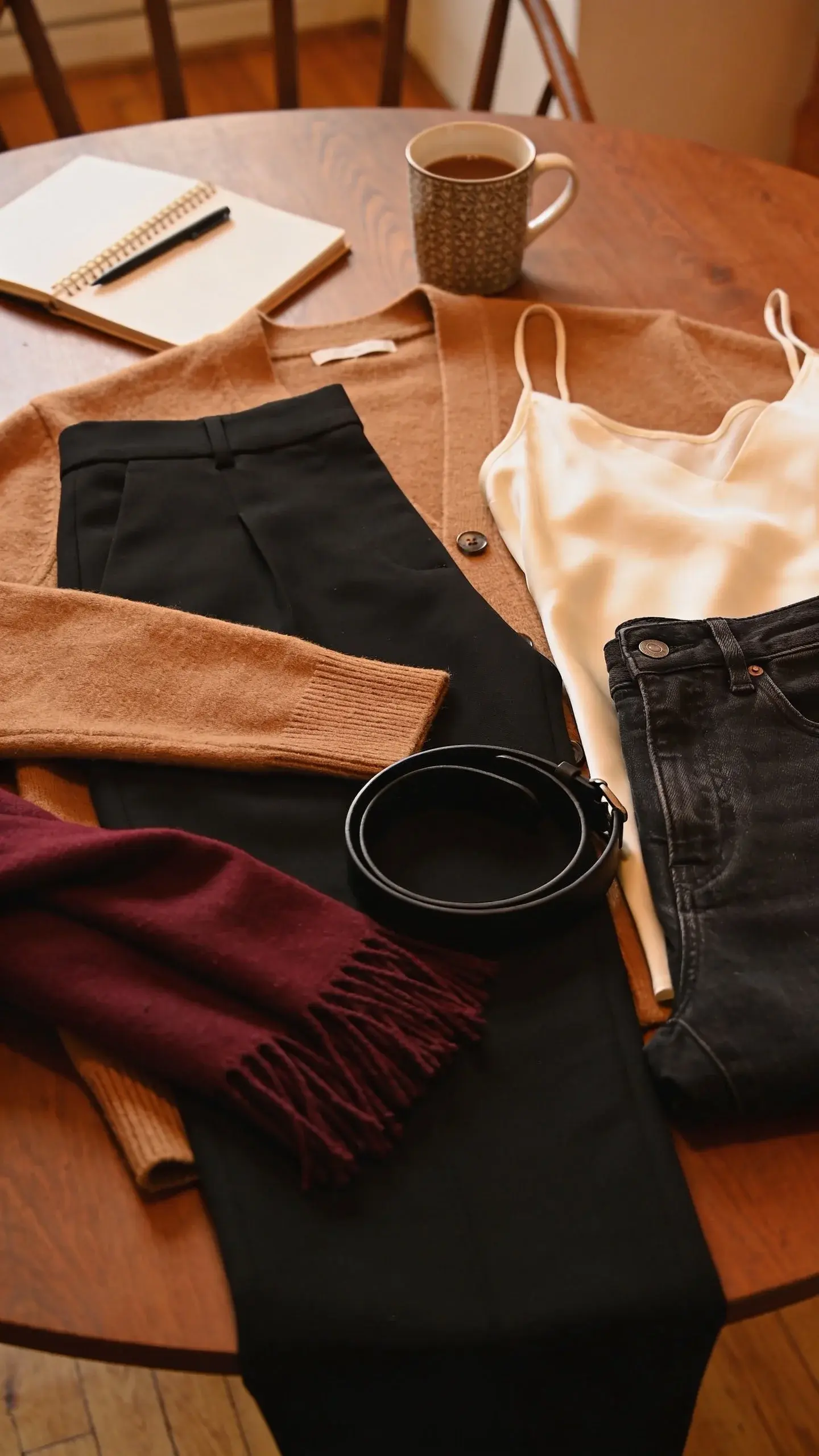Fall work-to-weekend mix on a wooden table: camel cardigan, black tailored trousers, cream silk camisole, dark denim, burgundy scarf, minimal black belt; coffee mug, notebook, and pen nearby to suggest versatility; warm amber lighting and rich textures.