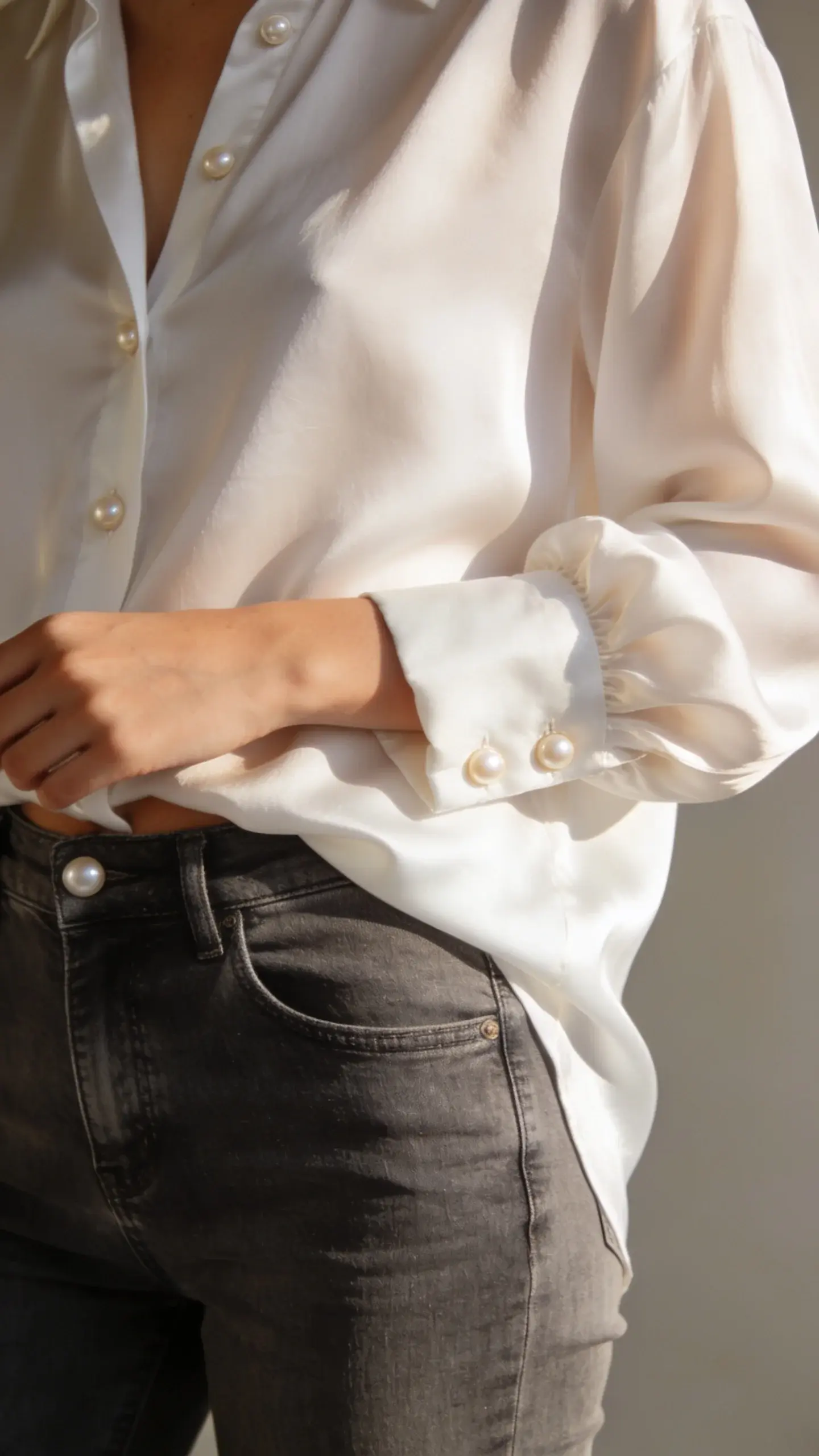 Close-up detail shot of luxurious fabric movement: a model’s torso in a fluid white cotton-silk shirt half-tucked into high-rise slim-straight jeans, sleeve slightly rolled, showcasing soft drape and pearly buttons; warm morning light, clean background, no text.