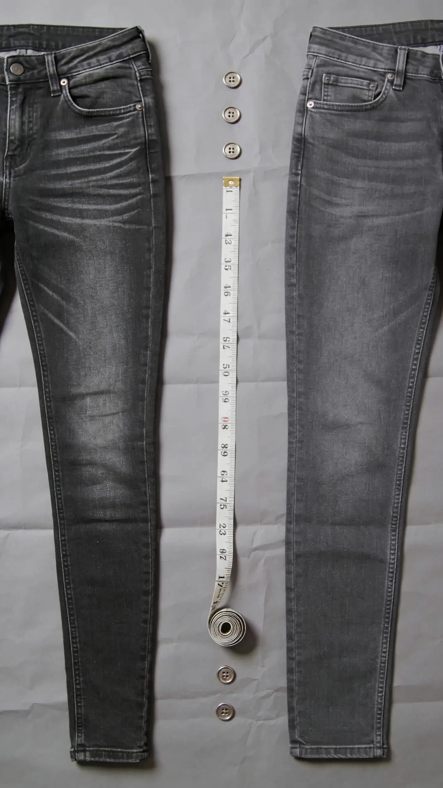 Before-and-after denim comparison flat lay on gray paper: left pair labeled by styling only (no text)—ultra-skinny jeggings with heavy whiskering; right pair—clean mid-wash slim-straight jeans with minimal hardware; include a measuring tape and metal buttons for context; neutral lighting, no text.