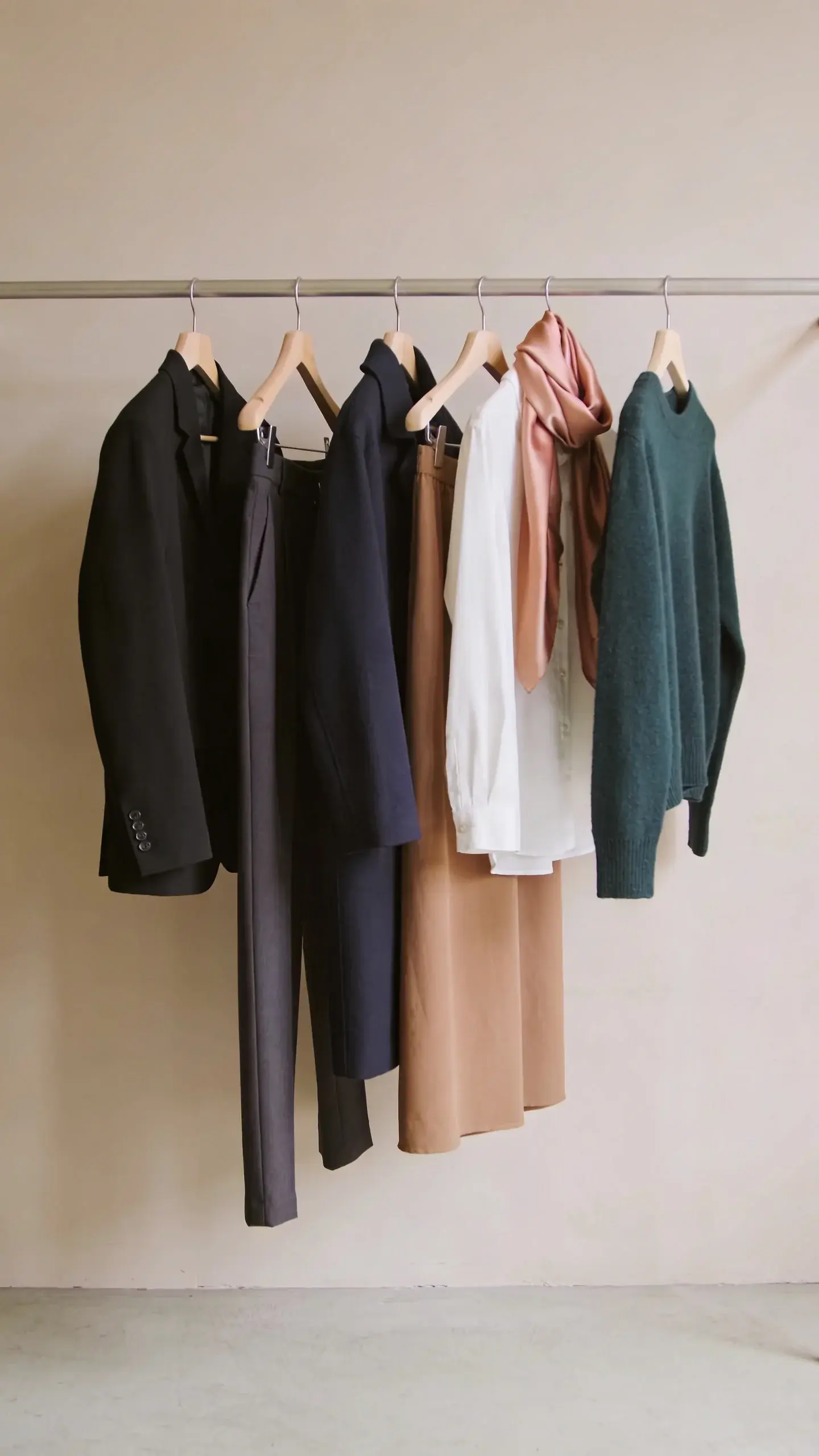 Capsule color story rack: neatly spaced hangers holding base neutrals (black blazer, charcoal trousers, navy coat, ivory blouse, taupe skirt) plus two accent pieces (soft blush silk scarf, deep forest green cashmere sweater); light beige wall, natural daylight, minimalist aesthetic.