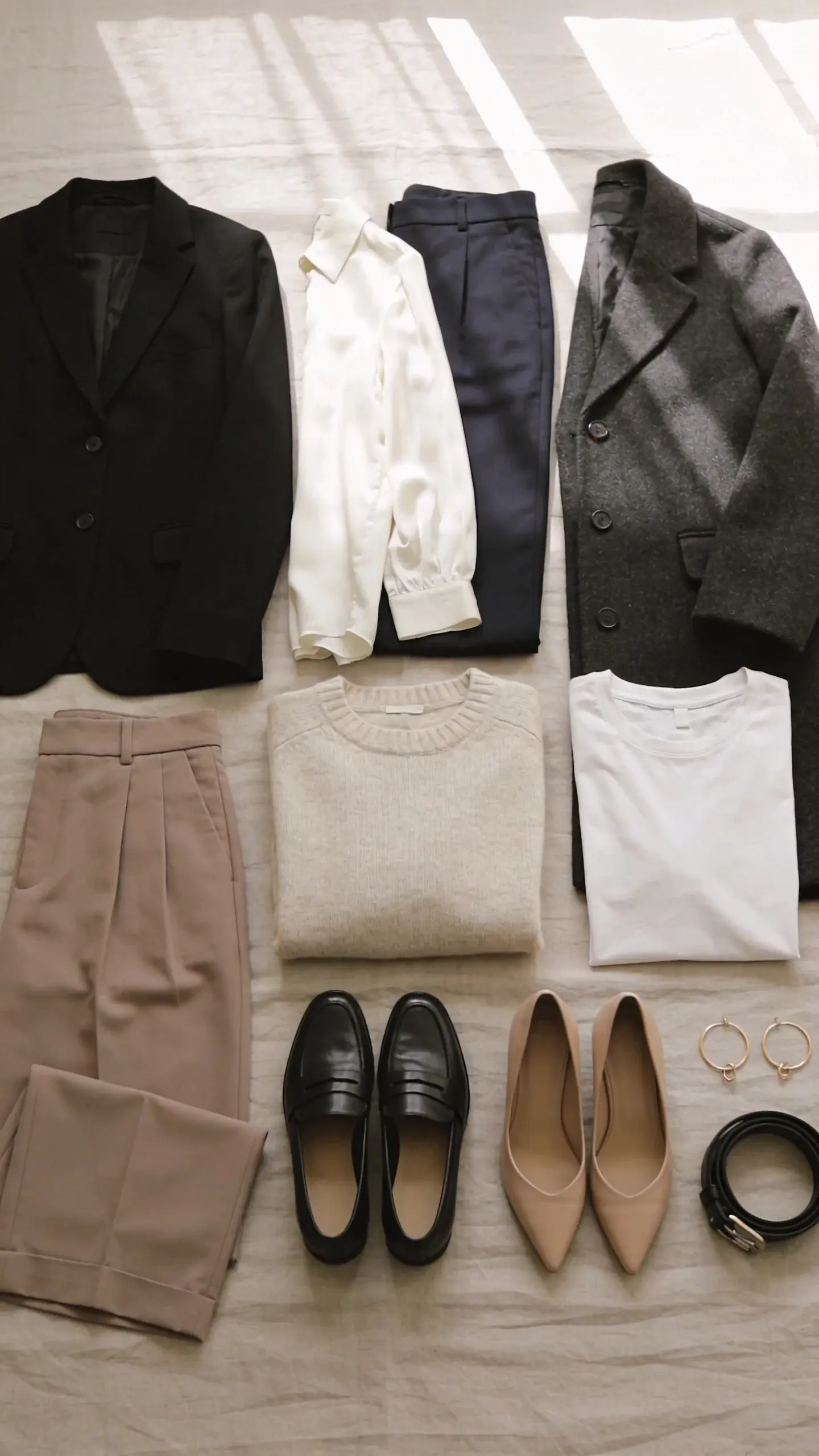 Flat lay of a neutral capsule wardrobe on a light taupe linen backdrop: tailored black blazer, deep navy trousers, charcoal wool coat, ivory silk blouse, oatmeal cashmere sweater, taupe wide-leg trousers, soft white tee, black leather loafers, nude pointed pumps, slim black belt, delicate gold hoop earrings; soft natural window light, minimal shadow, no text.