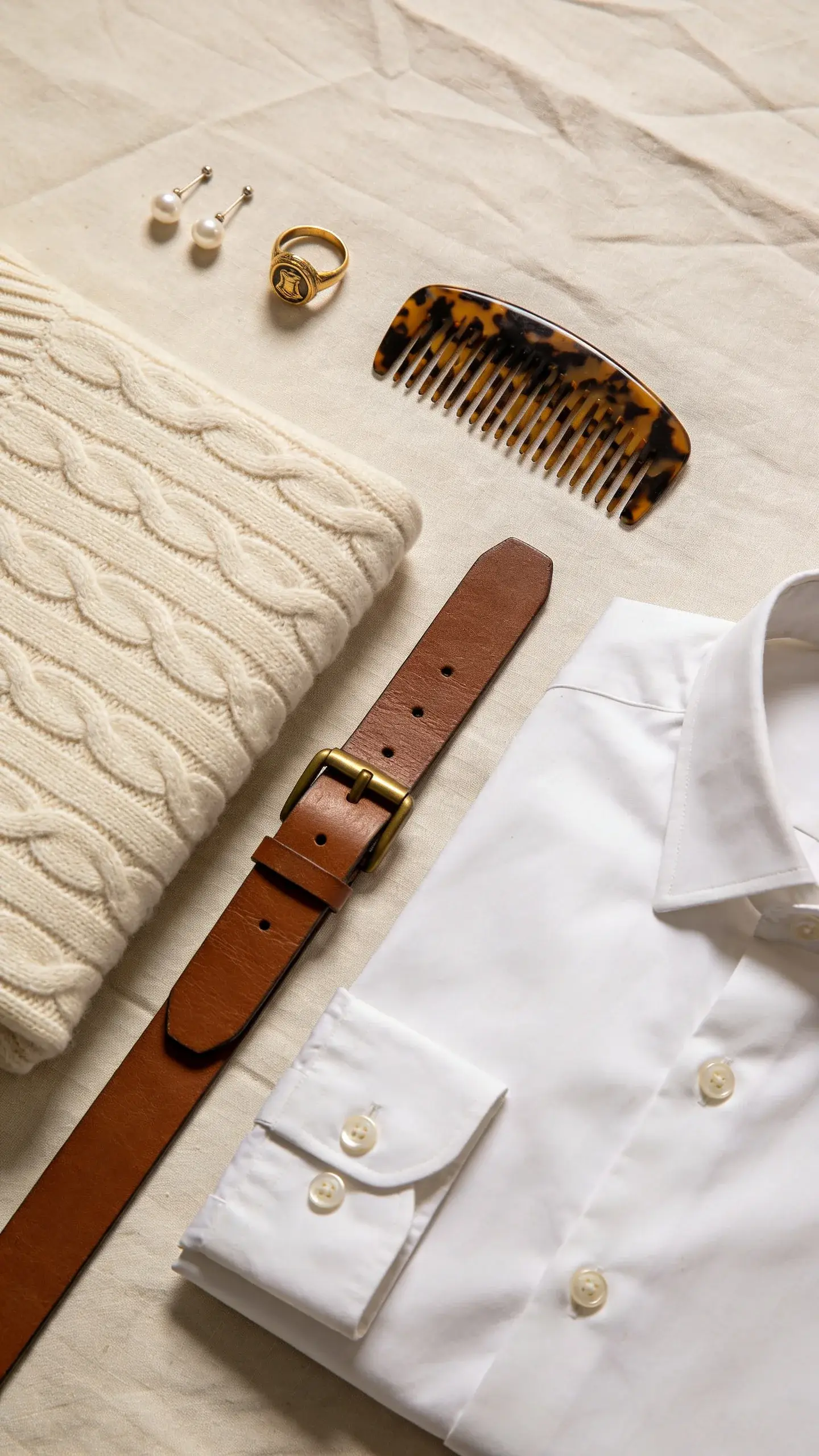 Close-up flat lay of Old Money essentials: pearl stud earrings, a vintage gold signet ring, tortoiseshell hair comb, neatly folded cream cable-knit cashmere sweater, crisp white oxford shirt with mother-of-pearl buttons, a slim brown leather belt with an unbranded brass buckle, all on a linen backdrop with gentle creases, neutral tones, no text.