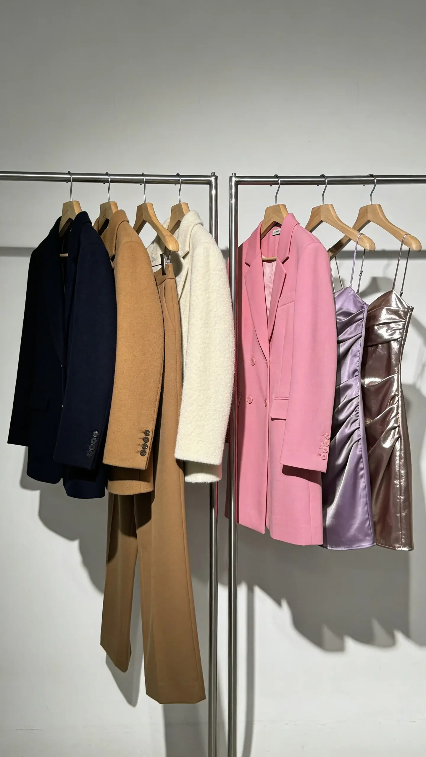 Side-by-side wardrobe rails split mood (no text, no labels): left rail with navy, camel, cream blazers and trousers in wool and cashmere on wooden hangers; right rail with bubblegum pink, lavender, and metallic mini dresses in satin and vinyl on chrome hangers; soft vs. high-gloss lighting contrast, clean studio backdrop.
