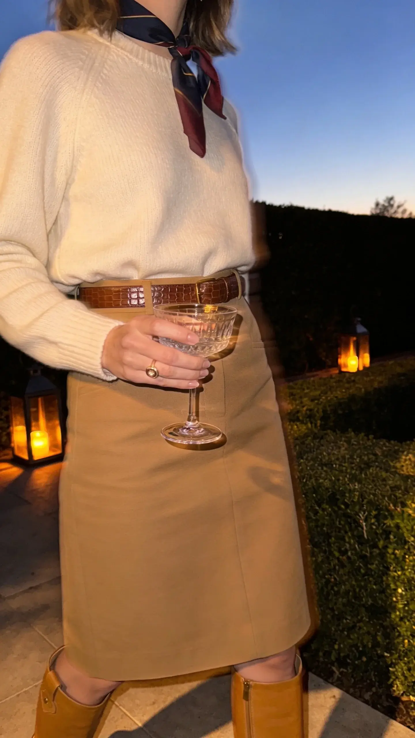 Country club terrace at dusk, woman in a cream cashmere crewneck tucked into a high-waisted camel A-line midi skirt, tan knee-high leather riding boots, thin silk scarf in muted navy/burgundy tied at neck, brown croc-embossed belt, delicate signet ring, holding a crystal coupe; manicured hedges and lantern glow in background.