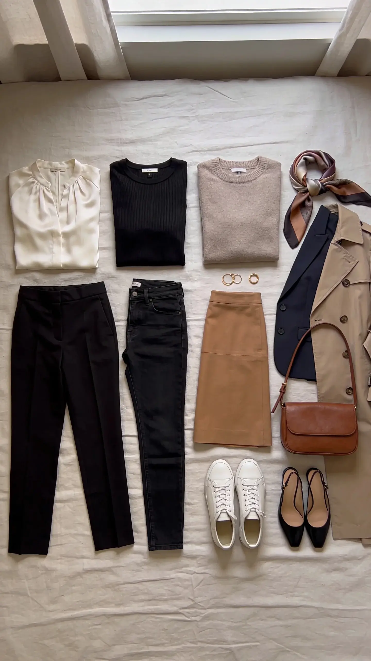 A neatly arranged flat lay of an elegant women’s travel capsule wardrobe on a light linen bedspread: two neutral tops (ivory silk blouse, black ribbed tee), one soft taupe sweater, tailored black trousers, slim dark jeans, a camel midi skirt, a lightweight navy blazer, a trench coat, a silk scarf in muted tones, minimalist gold jewelry, a leather crossbody bag, a pair of white leather sneakers, and low-heel black slingbacks; soft natural window light, no text.