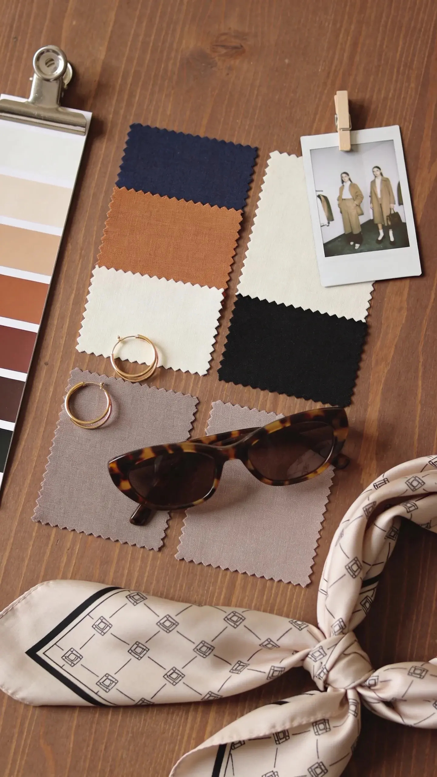A color strategy mood board on a wooden table: fabric swatches in navy, camel, ivory, black, and soft taupe; a color wheel clipped to the side; small accessories (gold hoop earrings, tortoiseshell sunglasses) and a silk scarf with subtle geometric print; a Polaroid of outfits pinned nearby; overhead natural light, no text.