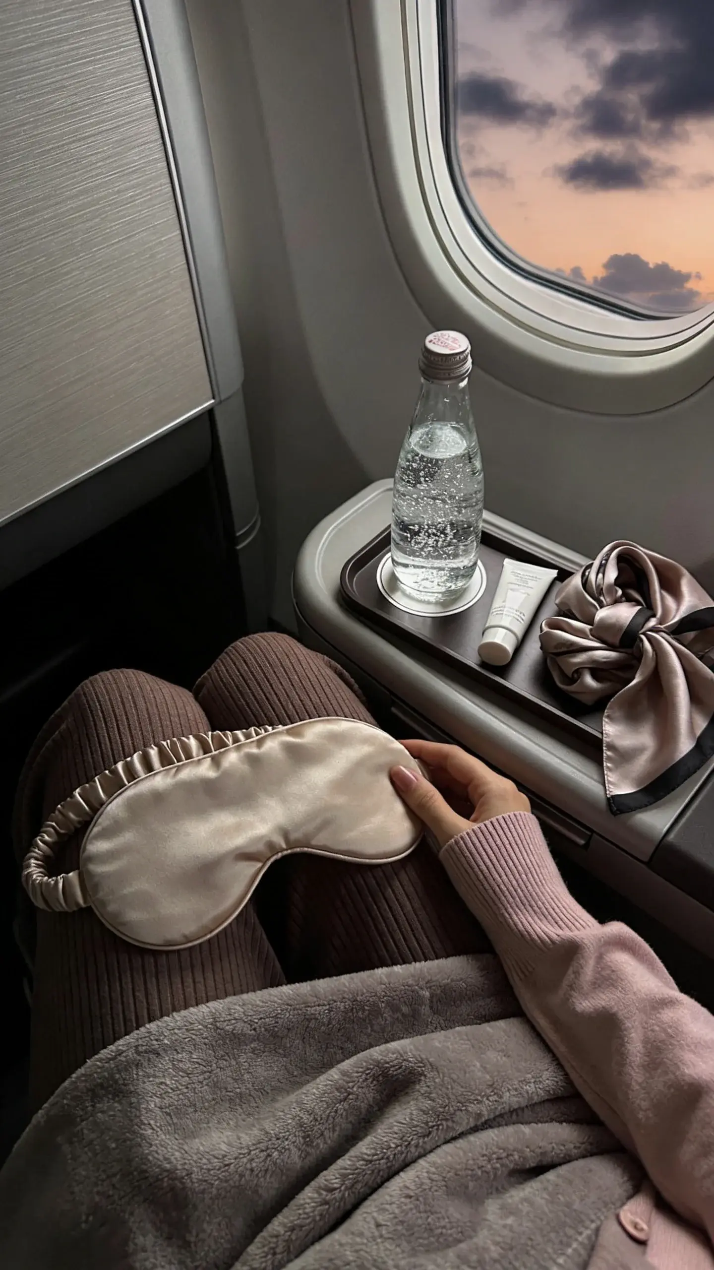 Window seat vignette: overhead view of a woman’s lap wearing mocha rib-knit trousers and a sand silk sleep mask pushed up to her forehead; pale blush cardigan sleeves; tray table with sparkling water, hand cream, and a neatly folded silk scarf; soft blanket in warm gray draped over knees; dusk clouds through the window, moody, no text.