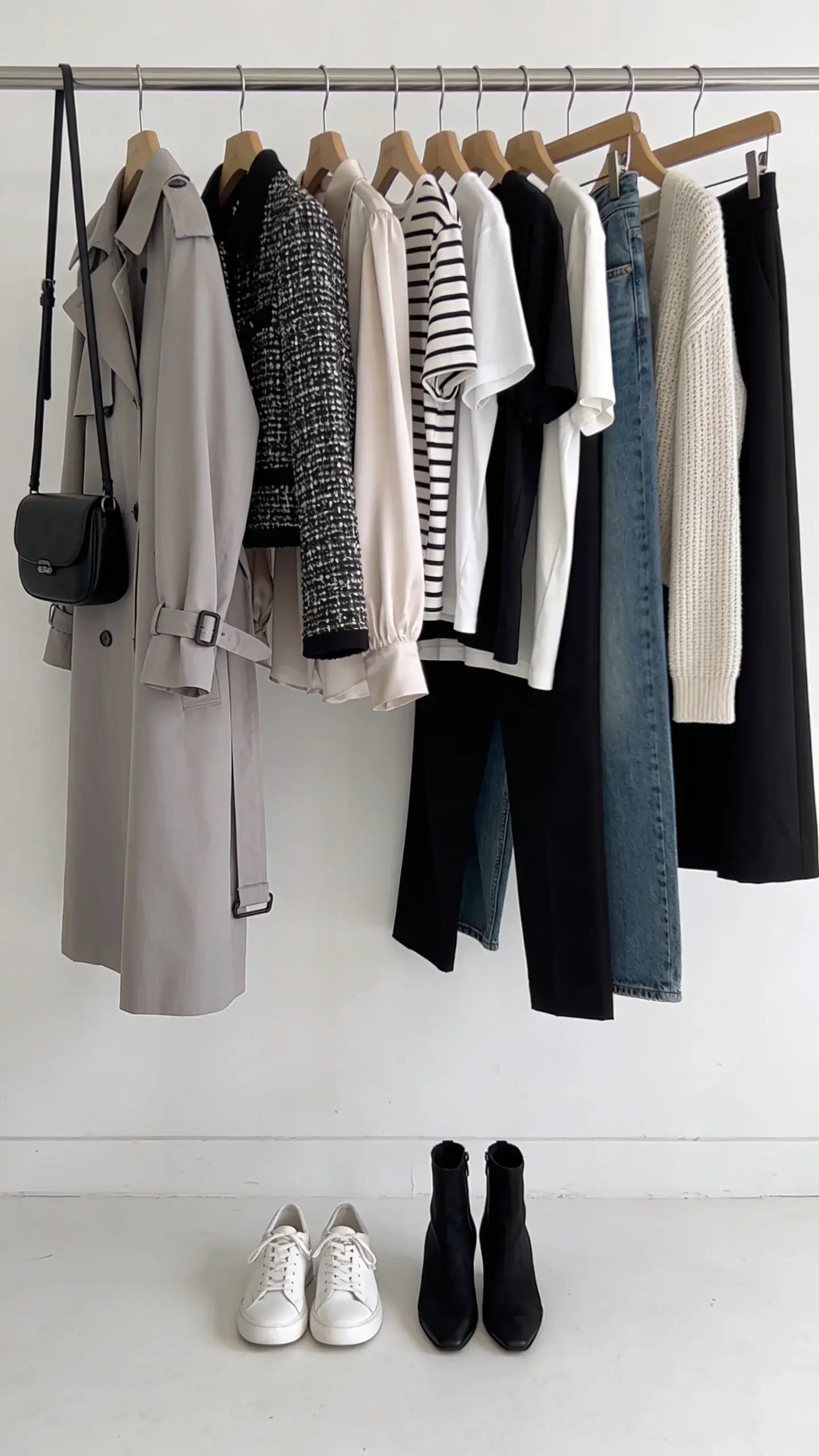A coordinated capsule wardrobe on a clothing rack against a white wall: trench, cropped tweed jacket in black-and-white, silk blouse, Breton stripe top, white and black tees, fine-knit cardigan, black trousers, medium-wash jeans, black midi skirt; below, white sneakers and black ankle boots aligned, and a compact black crossbody bag hanging on the rack.