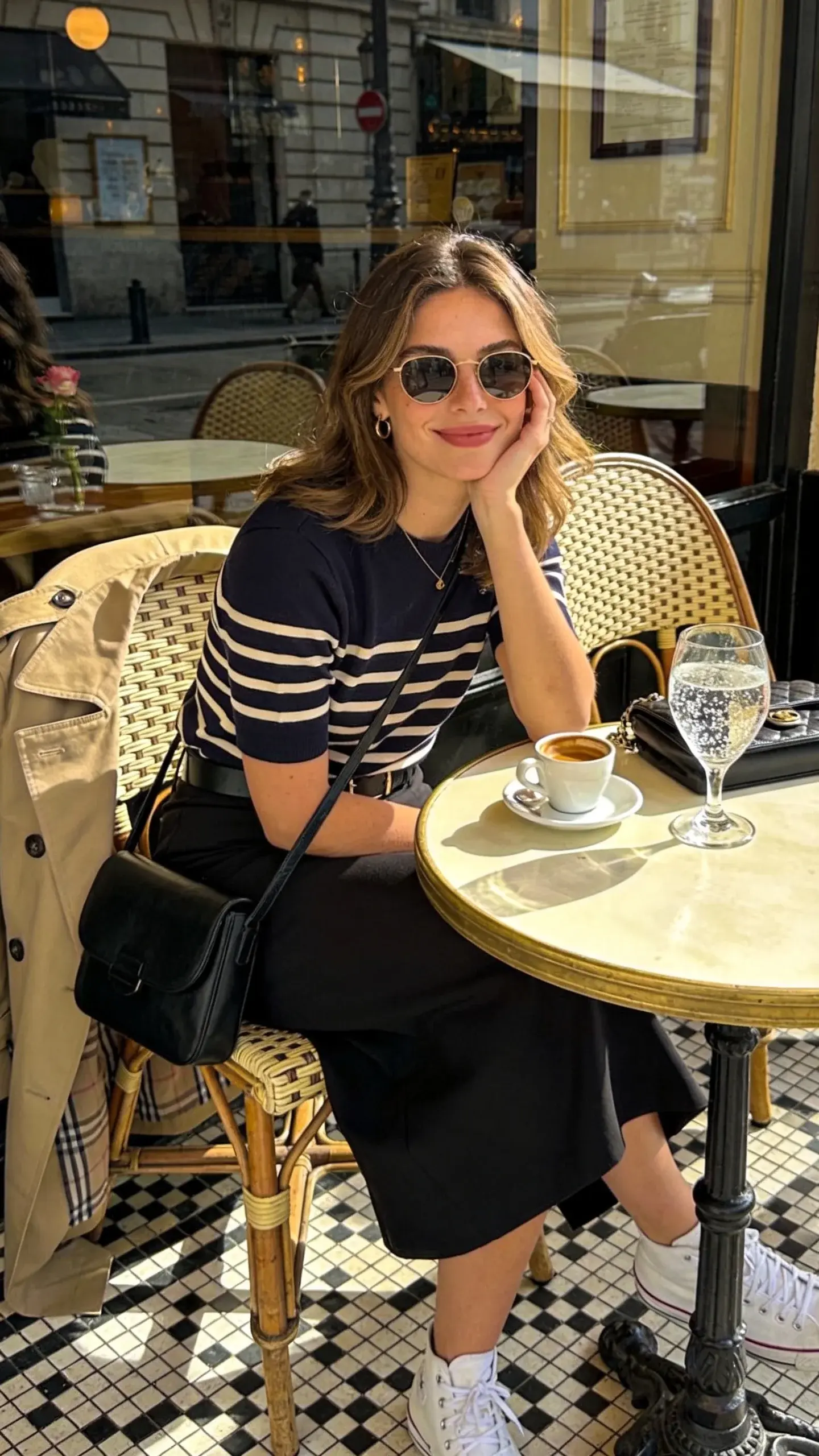A café patio scene at midday: a woman seated with a tiny espresso and sparkling water, wearing a navy-and-cream Breton top tucked into a black midi skirt, white sneakers, trench draped over the chair back, black crossbody bag on the table; wicker chairs, checkerboard tile floor, and street reflections in the café window.