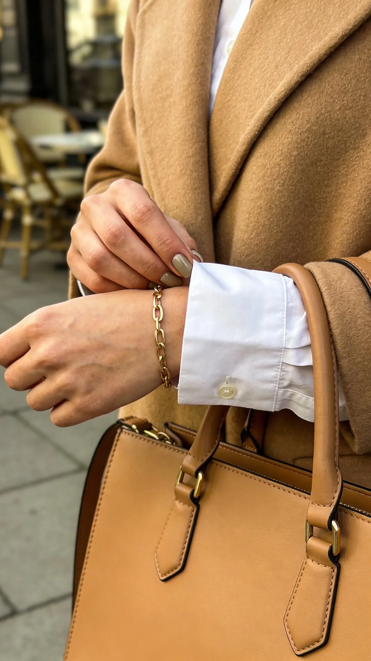 Detail shot outdoors: the woman’s hand adjusting a cuff on a crisp white shirt, layered under a camel coat; gold bracelet and subtle manicure; the strap of a structured leather tote visible; blurred city sidewalk and café chairs in the background—no text.
