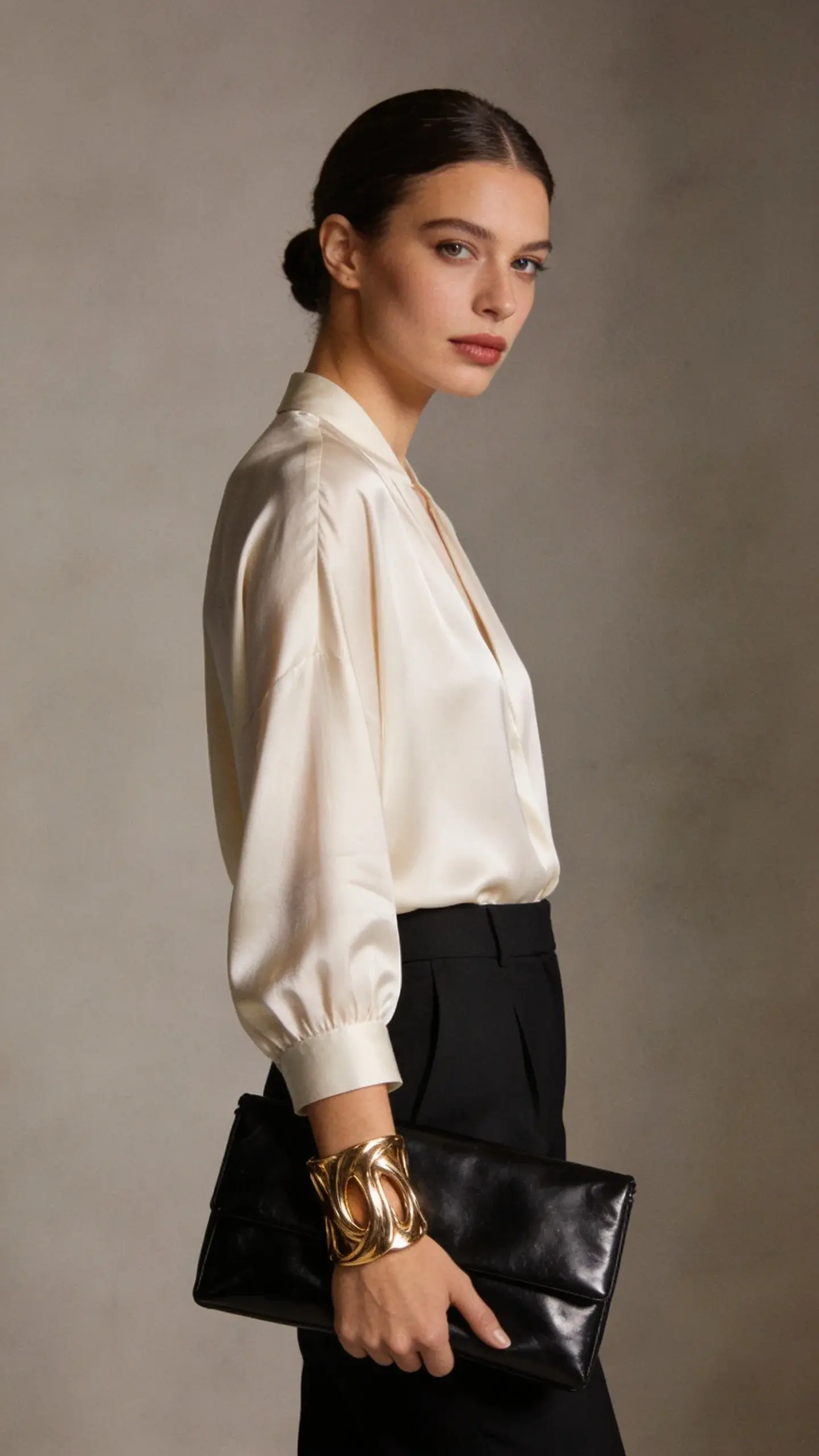 A minimalist, softly lit studio portrait of an elegant woman in a cream silk blouse and tailored black trousers, wearing one bold sculptural gold cuff bracelet; no visible logos; smooth low bun, natural makeup; holding a simple black polished leather clutch; neutral background in warm gray.