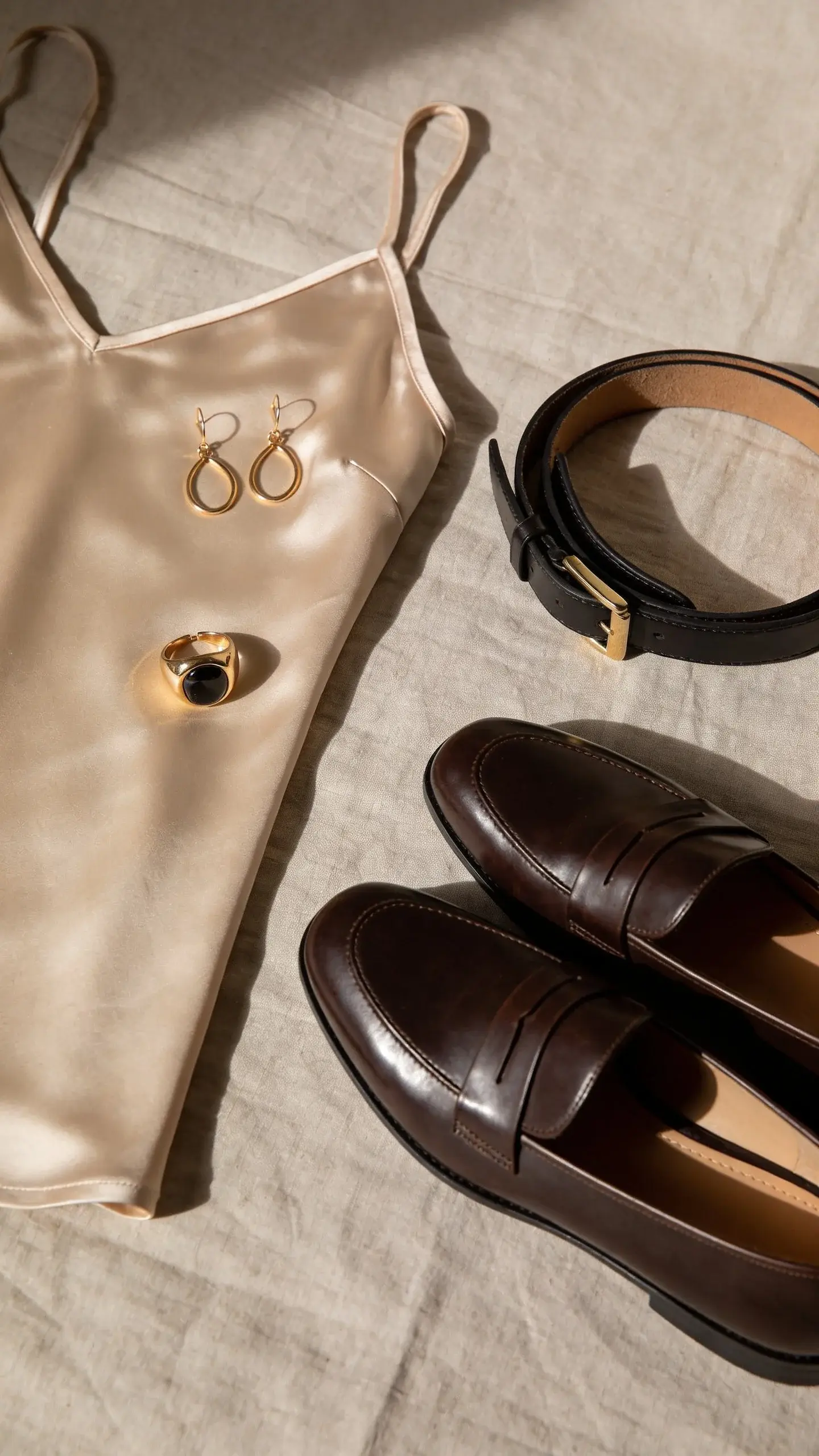 Close-up flat lay on a linen surface: satin camisole in champagne, minimalist gold earrings, a single bold statement ring, polished leather loafers, and a slim belt with an understated buckle; no brand names; soft shadows, neutral tones.