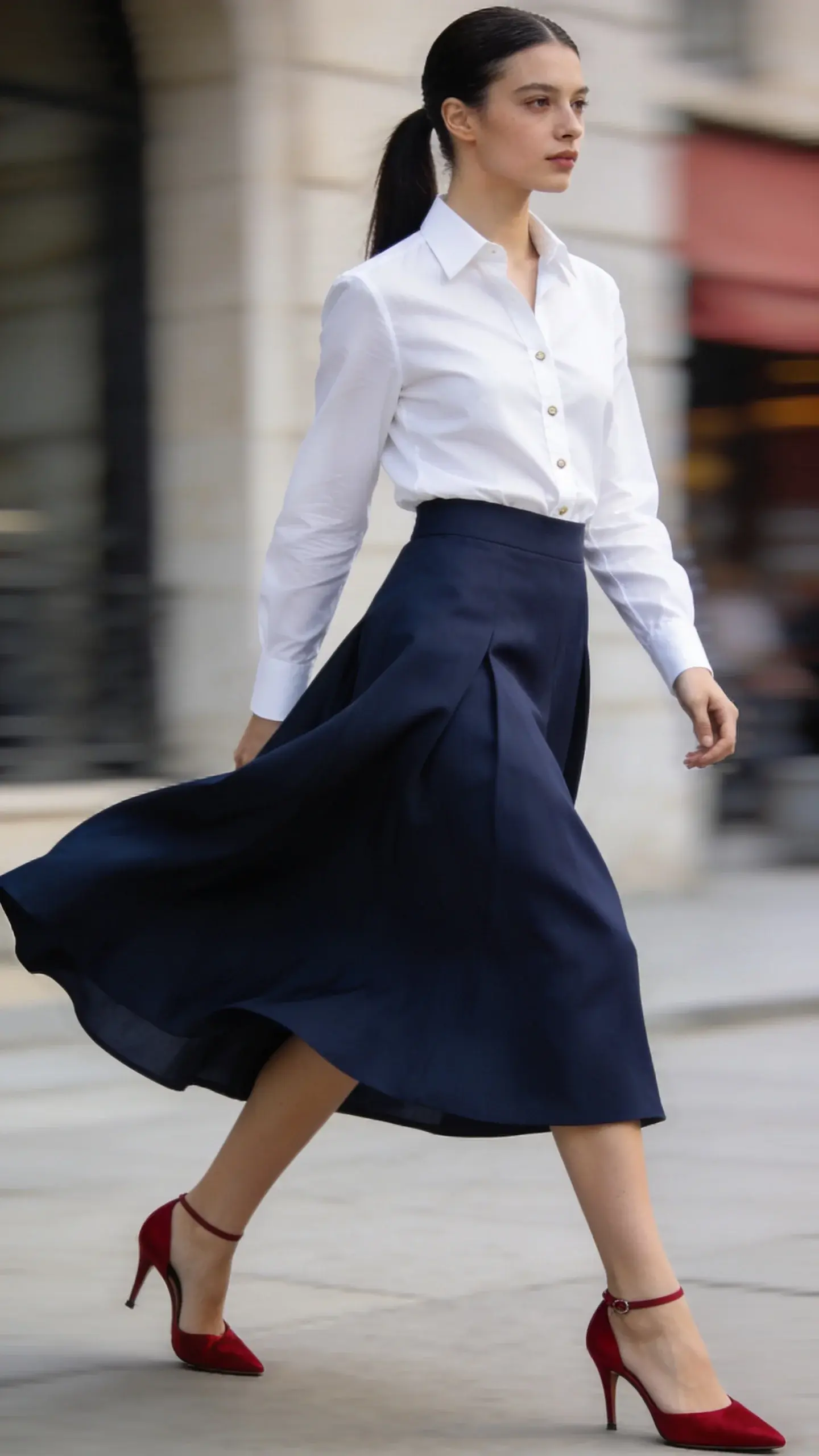 Editorial shot of a sweeping navy midi skirt moving in a gentle breeze, paired with a crisp white button-down neatly tucked; bare neckline, hair in a sleek ponytail; one standout item: cherry-red kitten-heel slingbacks; urban background softly out of focus.