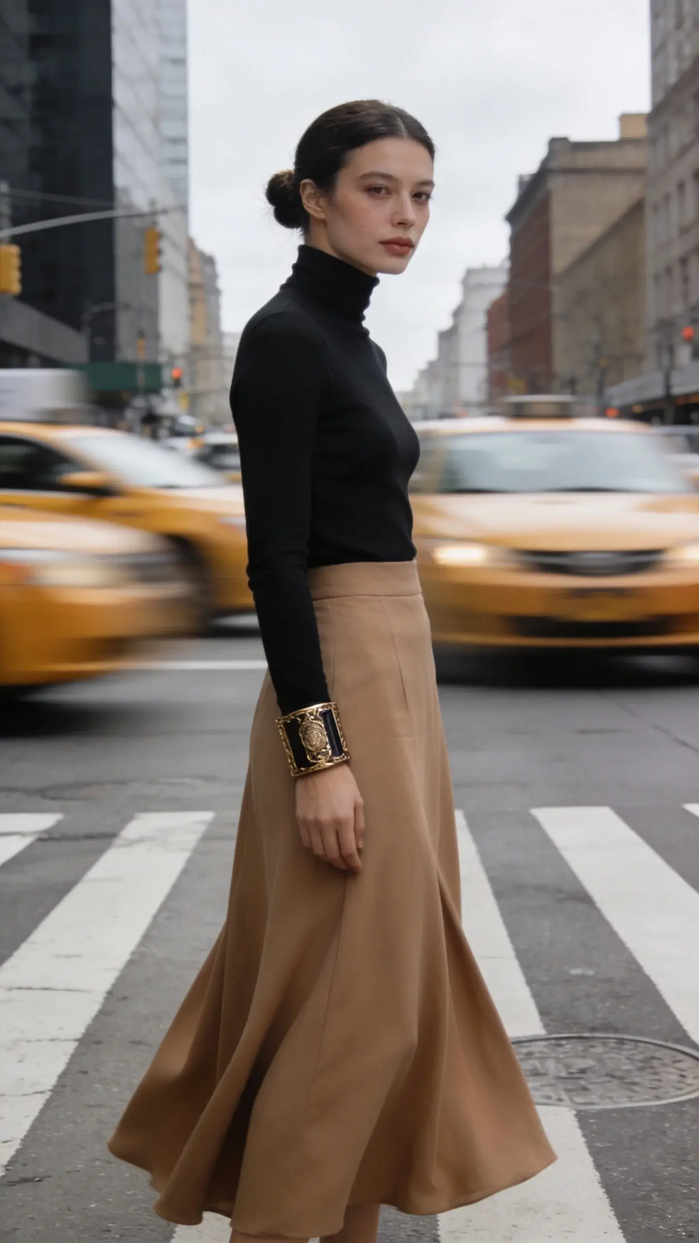 City crosswalk capture: woman wearing a black fitted turtleneck and a camel sweeping midi skirt; one focal accessory—bold cuff bracelet; hair tucked into a low knot; no flashy elements, neutral makeup; soft overcast daylight, blurred taxis behind.
